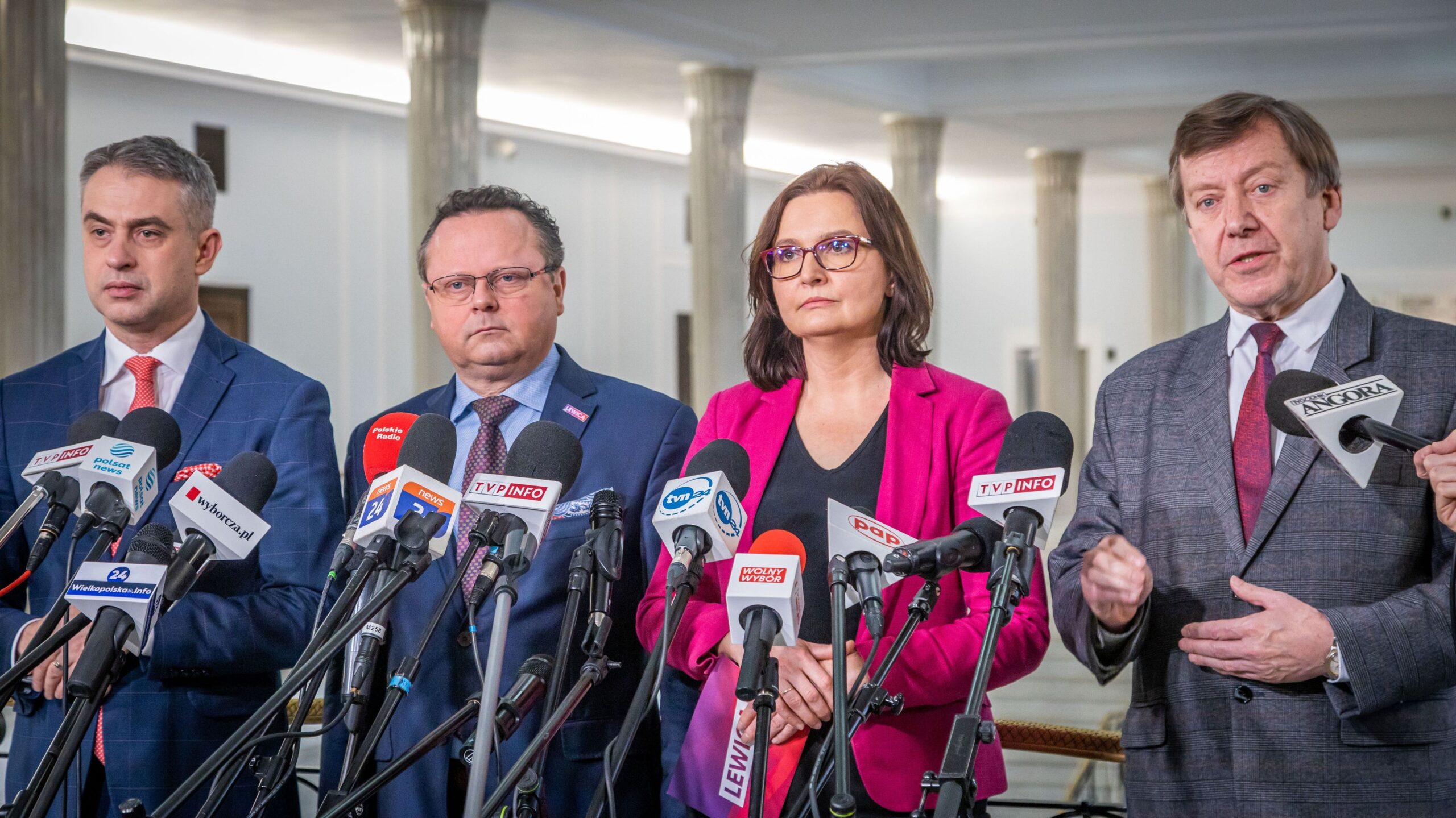 Czterech polityków stoi przed mikrofonami podczas konferencji prasowej w jasno oświetlonym wnętrzu.