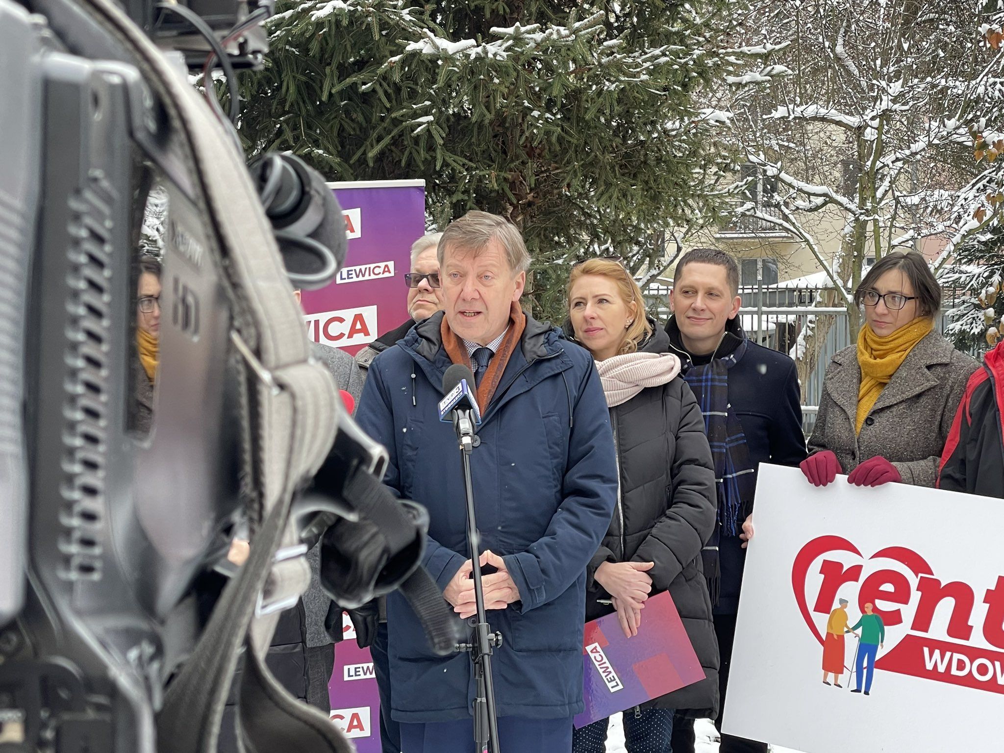 Na zdjęciu przedstawiono mężczyznę przemawiającego do mikrofonu podczas konferencji prasowej, z grupą osób w tle, w zimowej scenerii z zaśnieżonymi drzewami.