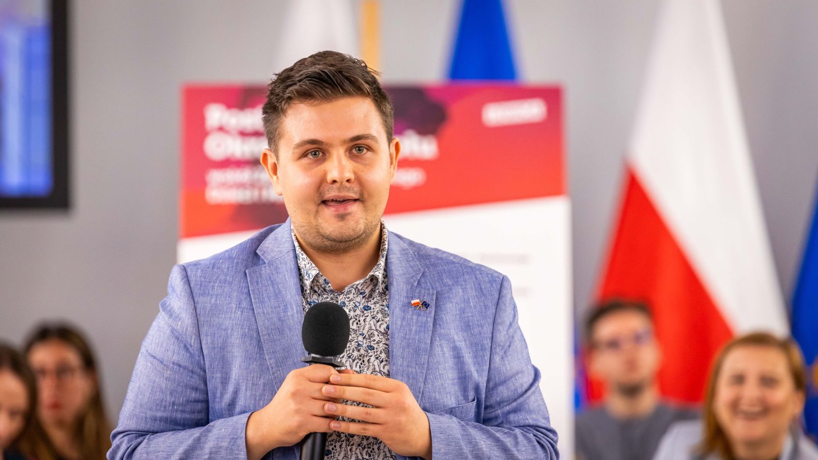 Mężczyzna w błękitnym garniturze trzyma mikrofon i przemawia do publiczności podczas podsumowania Okrągłych Stołów wokół zdrowia psychicznego dzieci i młodzieży w Polsce, z flagami w tle.