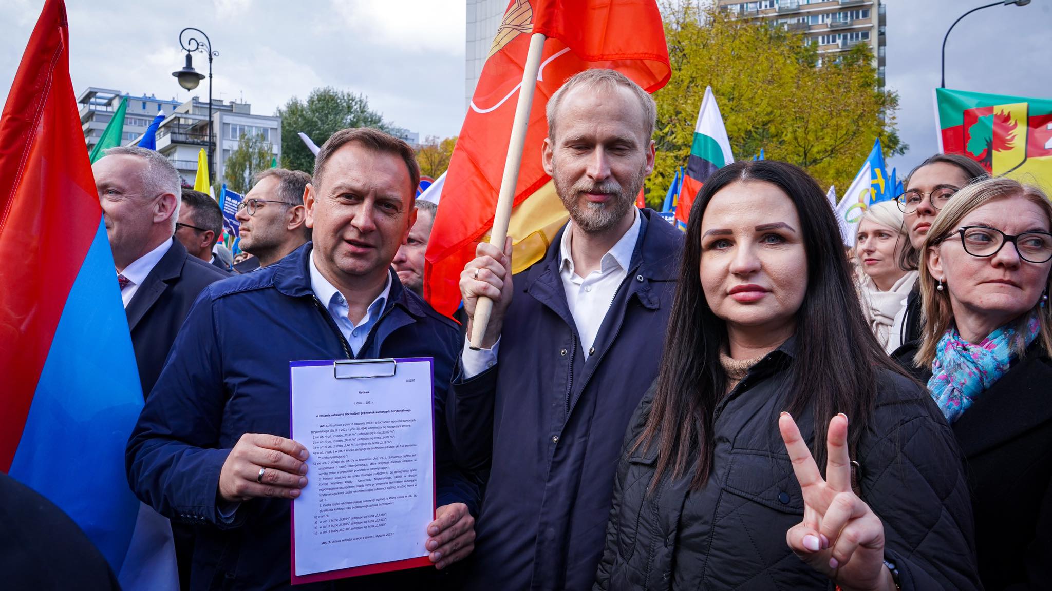 Grupa uczestników demonstracji w mieście niosących flagi i dokument w teczce, w obronie lokalnych społeczności i samorządności.