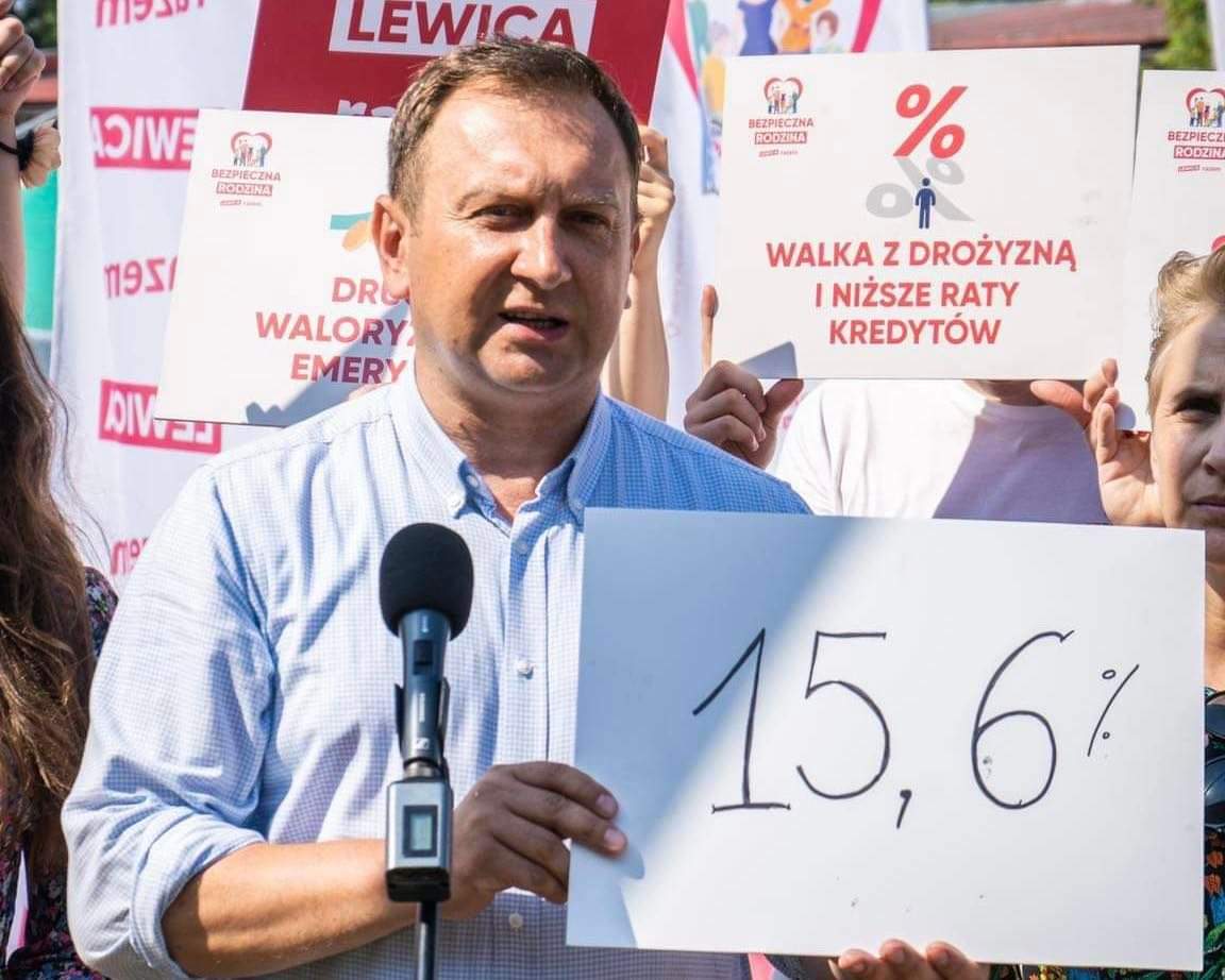 Mężczyzna stojący przy mikrofonie trzyma kartkę z wartością 15,6% podczas protestu przeciw inflacji, wokół widoczne są plakaty dotyczące drożyzny i obniżenia rat kredytów.