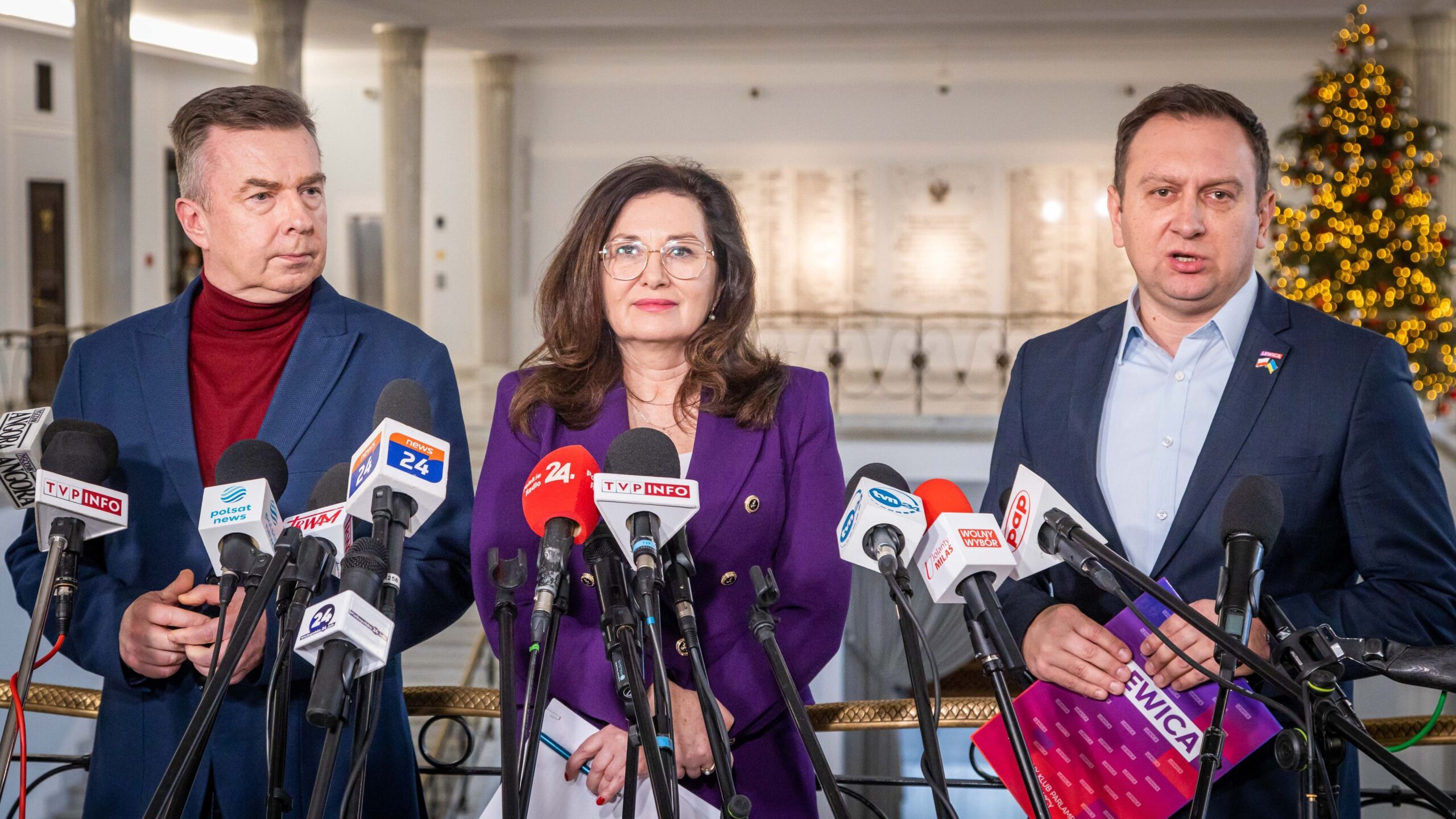 Na zdjęciu przedstawiono trzech polityków podczas konferencji prasowej w stylowym wnętrzu z choinką w tle.