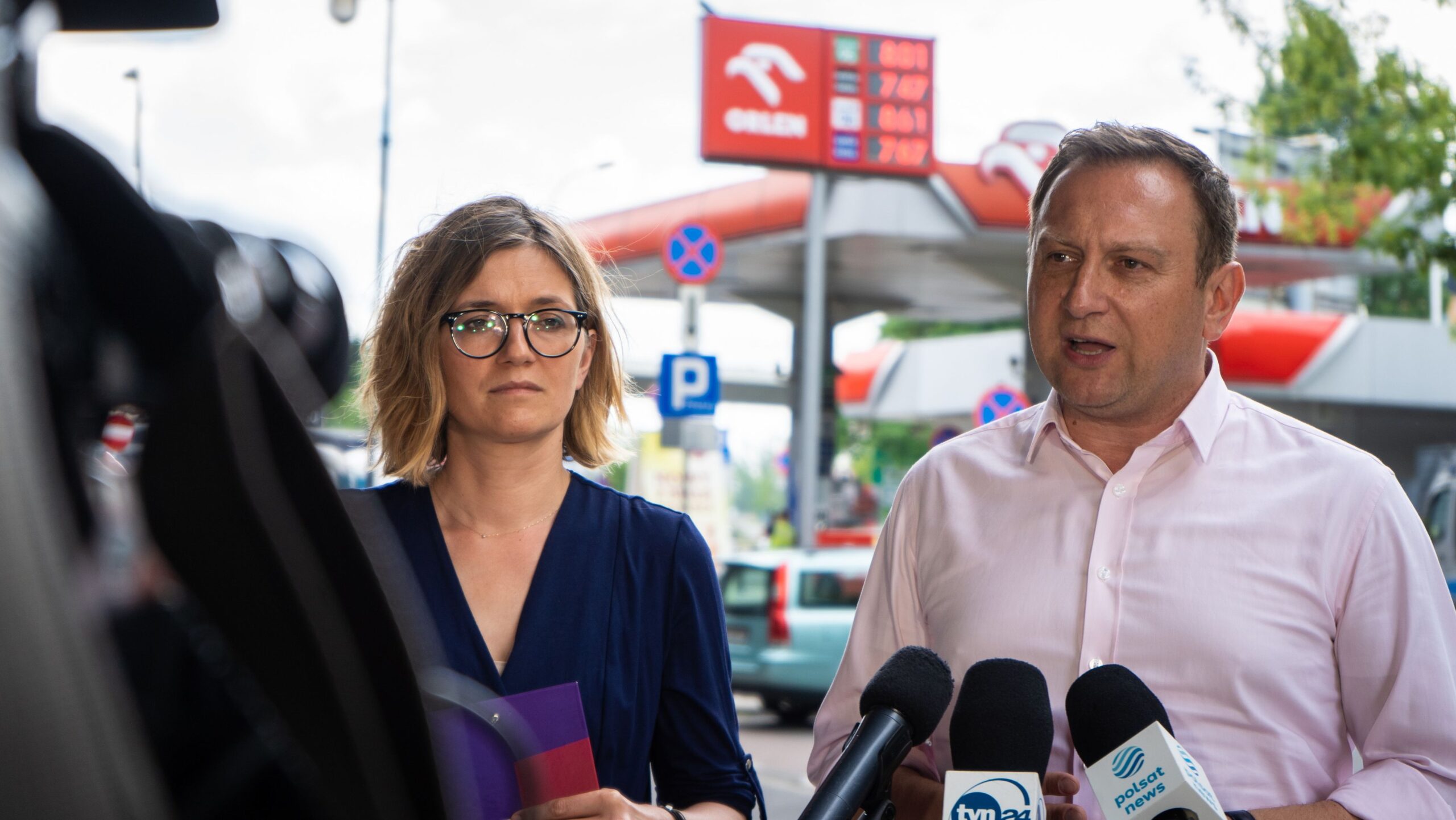 Na zdjęciu представione są dwoje ludzi podczas konferencji prasowej przed stacją, gdzie w tle widać oznaczenia cen paliw i znaki drogowe.