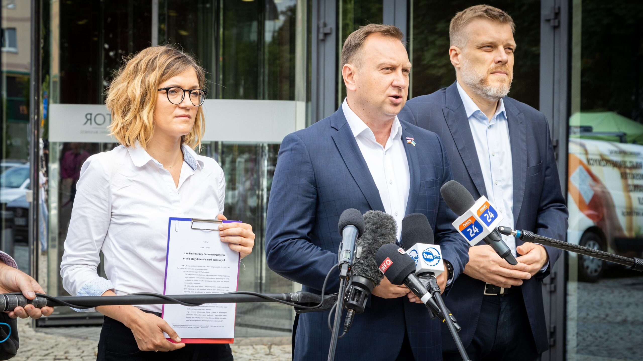 Konferencja prasowa, na której występują trzy osoby, z mikrofonami w ręku, w tle szklany budynek.