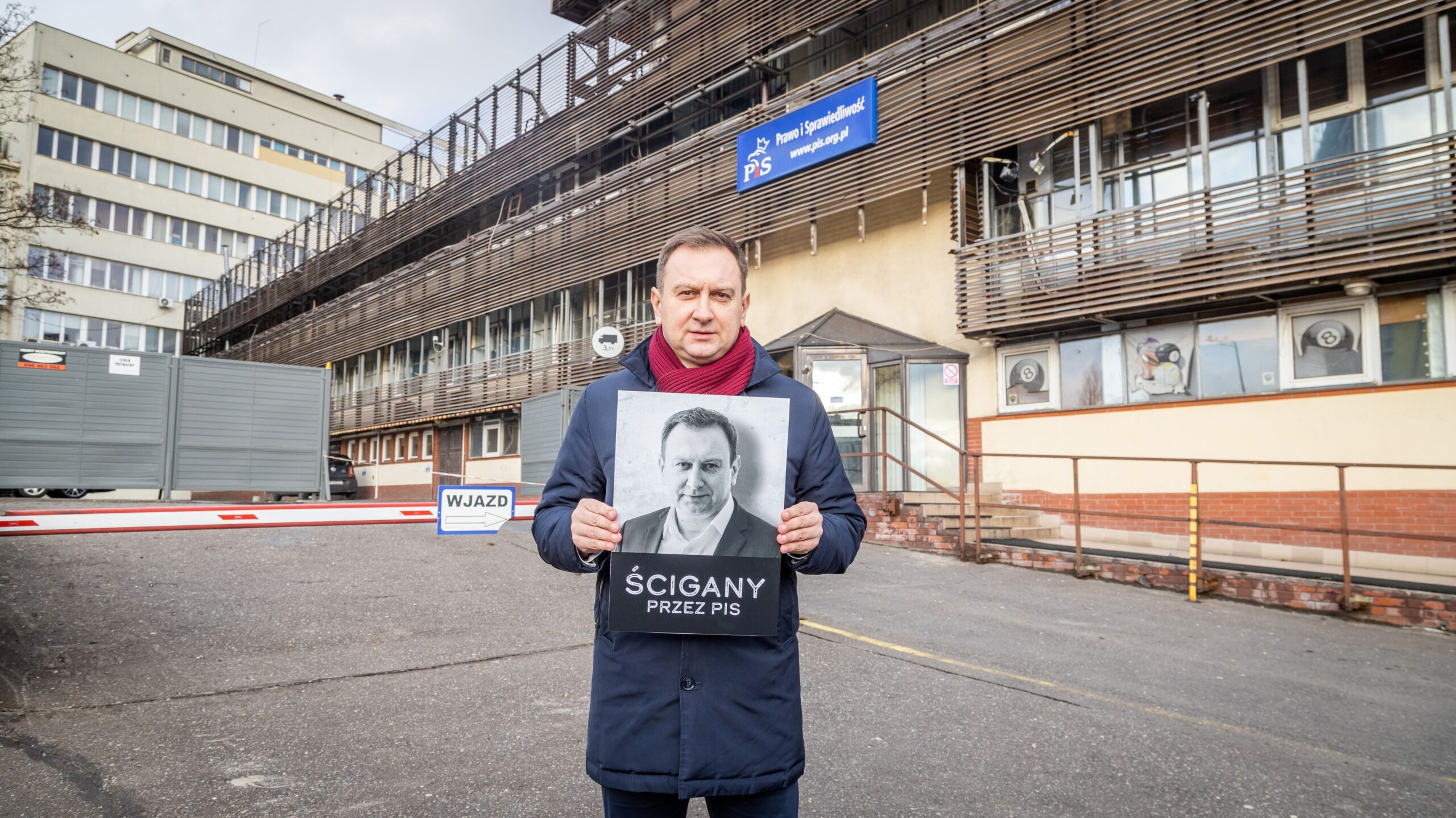 Mężczyzna stoi przed siedzibą partii politycznej z plakatem informującym o prześladowaniu w związku z działalnością Prawa i Sprawiedliwości.