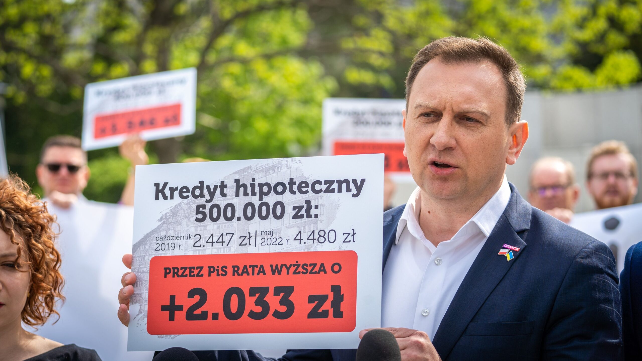 Mężczyzna w garniturze przemawia podczas demonstracji, trzymając tablicę z informacją o kredycie hipotecznym i rosnących ratach.