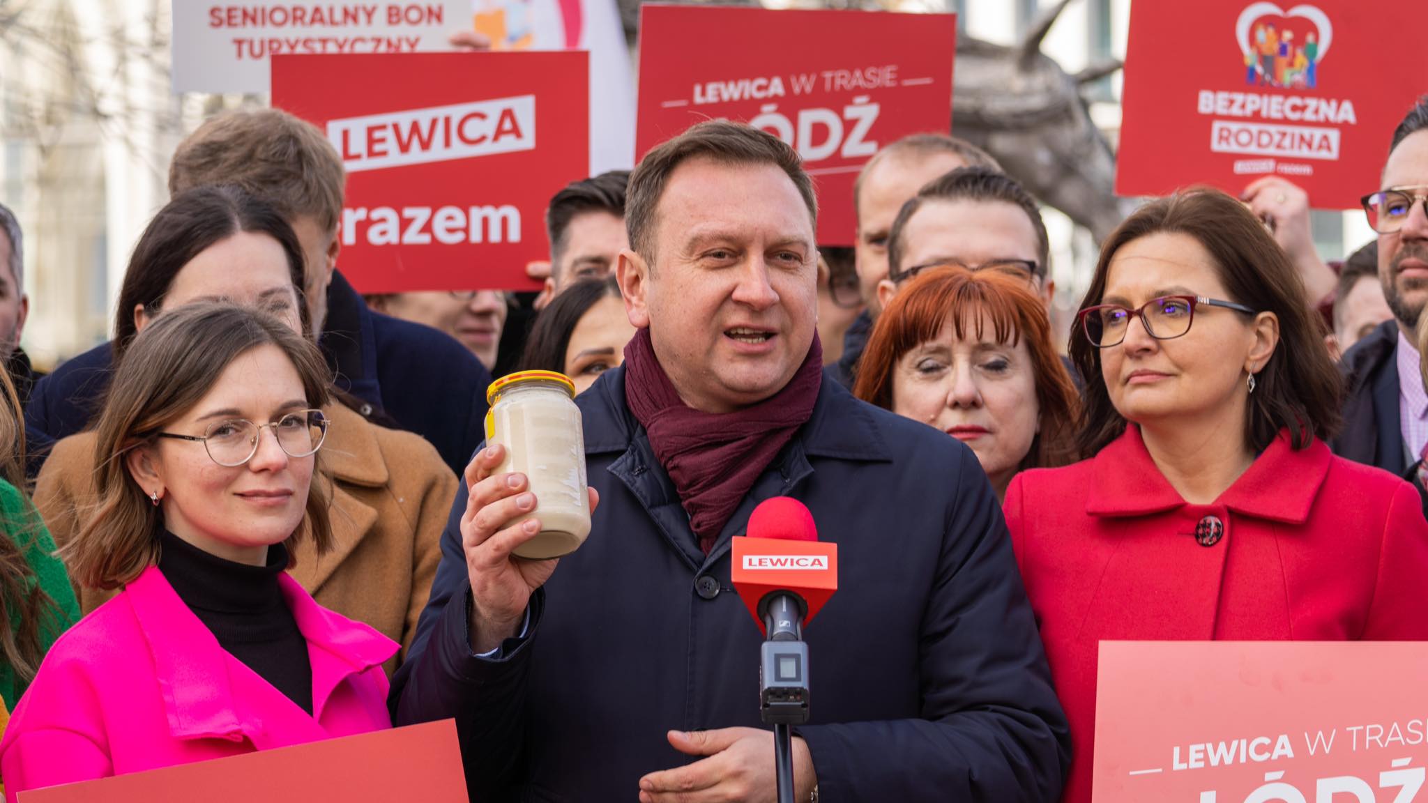 Na zdjęciu widać mężczyznę przemawiającego do zgromadzenia, trzymającego słoik, otoczonego przez grupę osób, które trzymają czerwone plakaty związane z kampanią Lewicy.