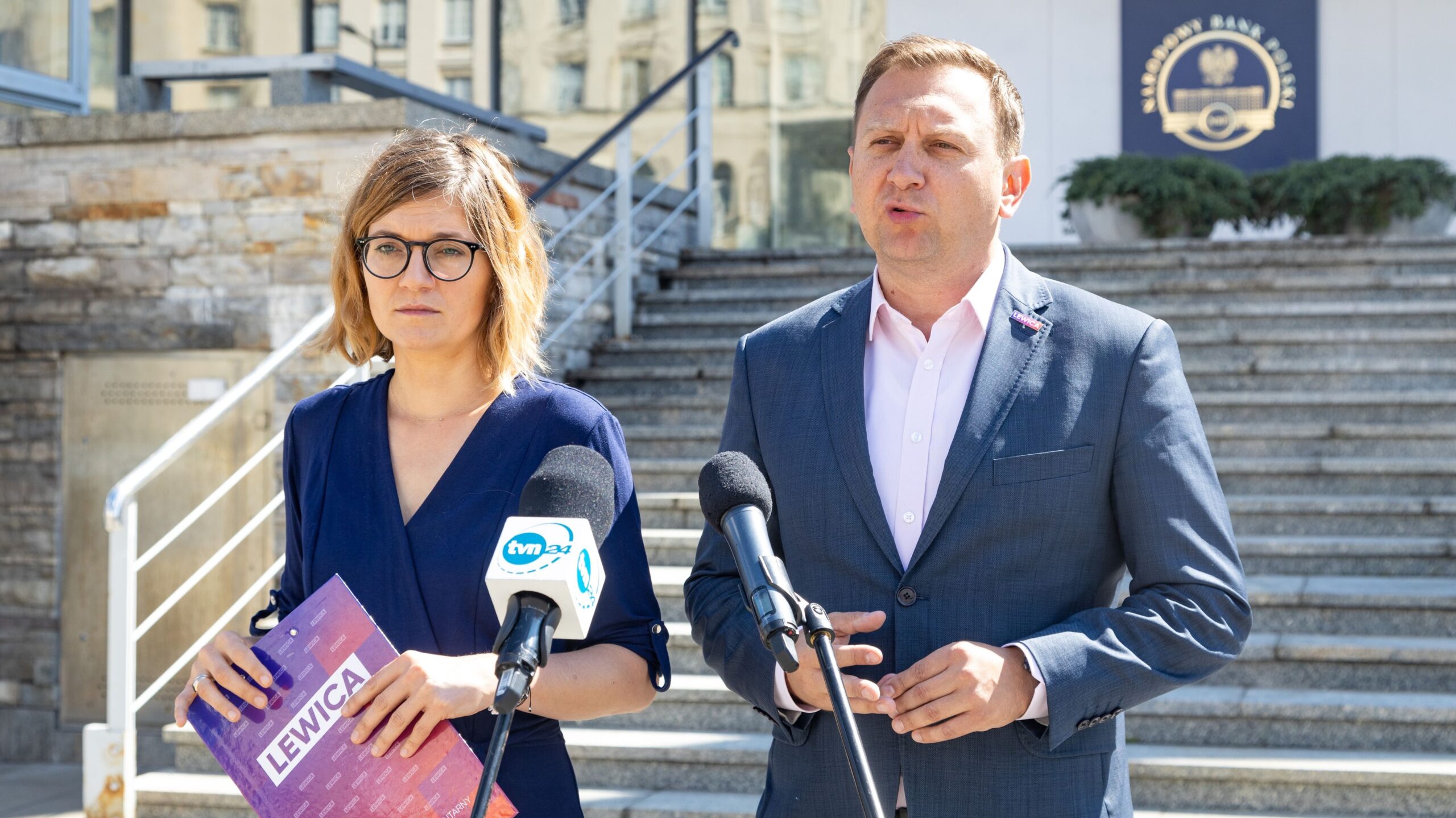 Dwoje polityków stojących na schodach przed budynkiem Narodowego Banku Polskiego, kobieta trzyma teczkę z napisem Lewica, a mężczyzna w garniturze wygłasza oświadczenie do mediów.