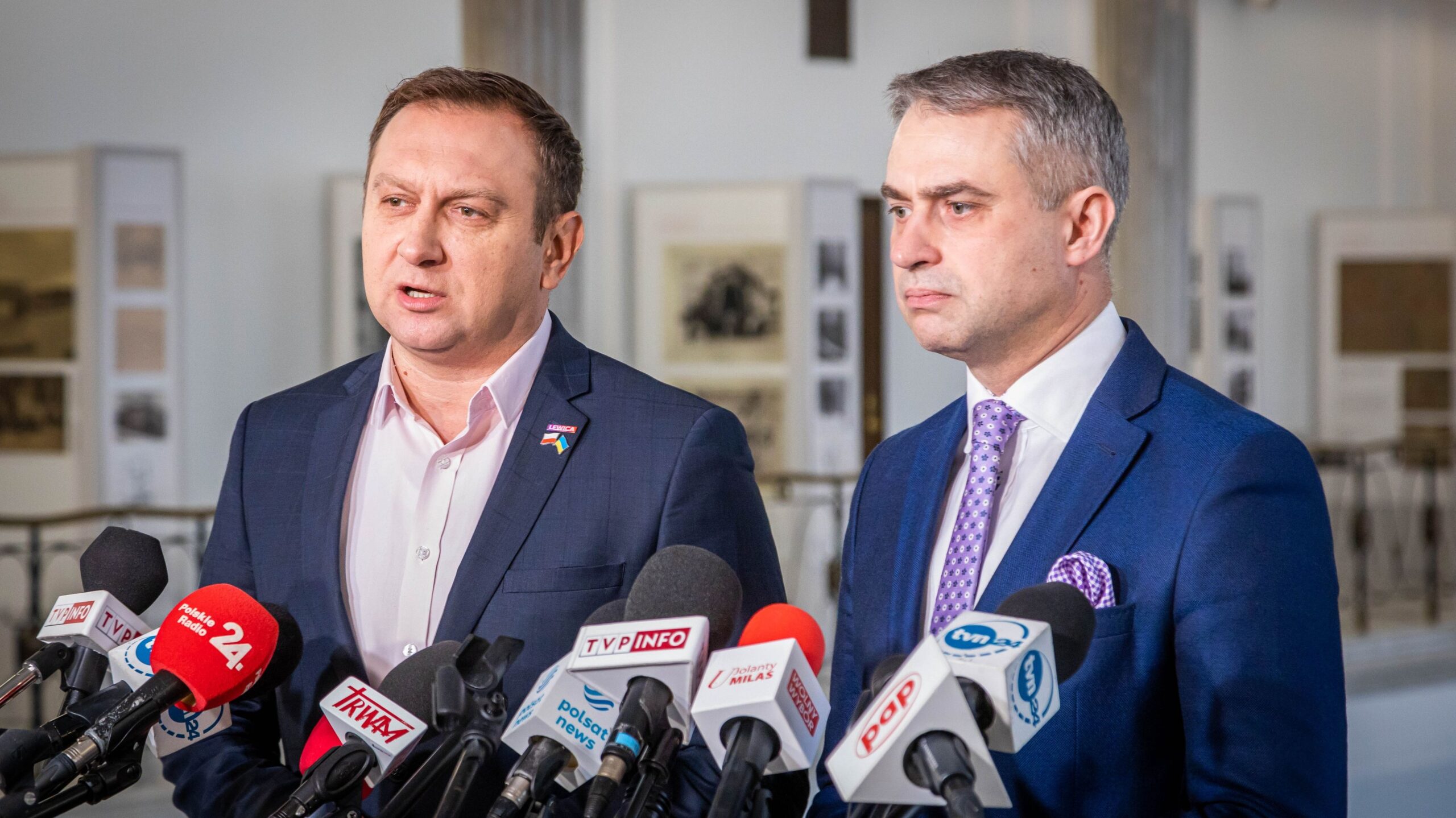 Na zdjęciu dwóch mężczyzn stoi przy mikrofonach i udziela wywiadu w tle wystawy.