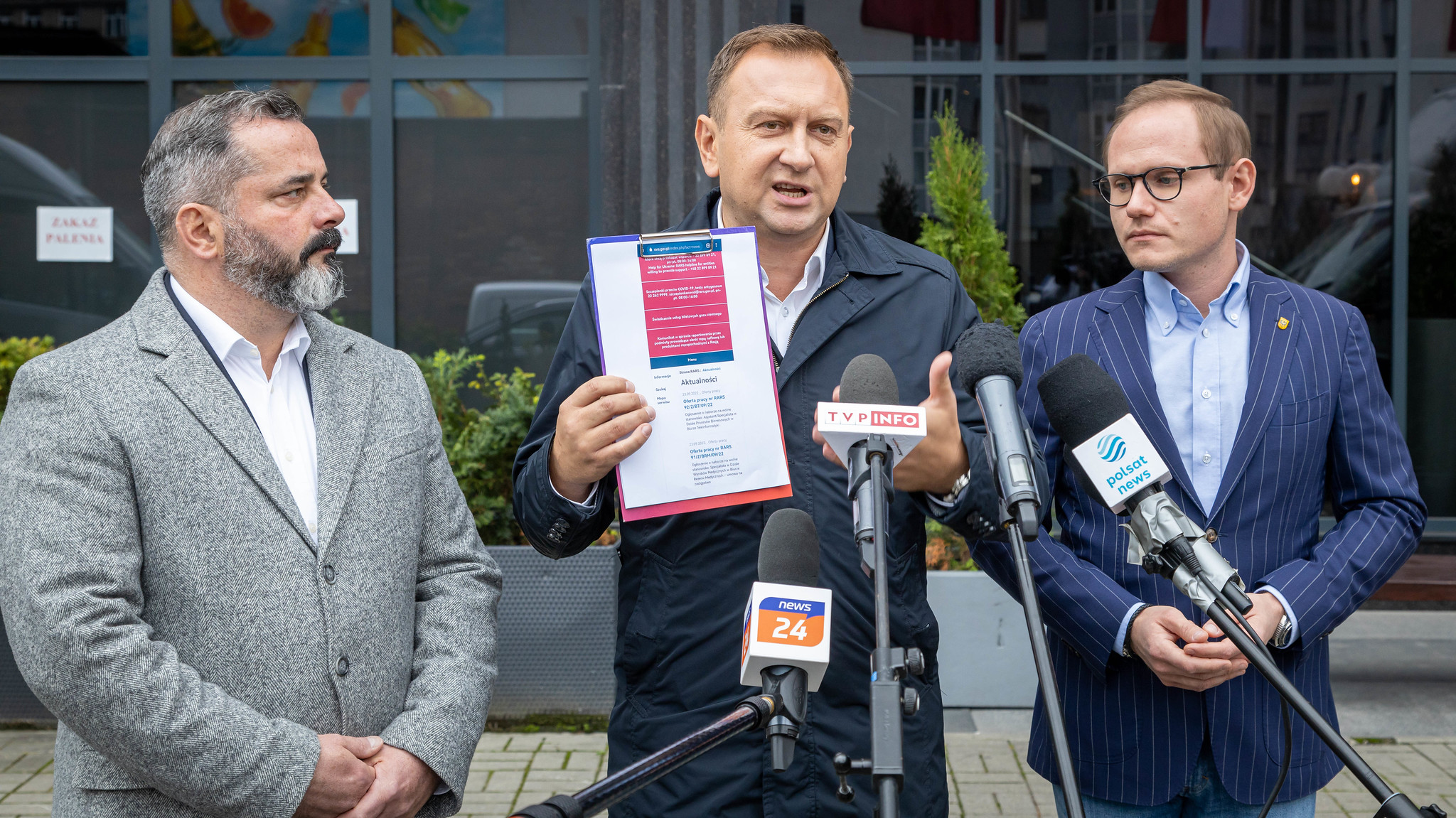 Na zdjęciu widać trzech mężczyzn podczas konferencji prasowej, przy mównicy z mikrofonami, gdzie jeden z nich trzyma dokument, a pozostali słuchają.