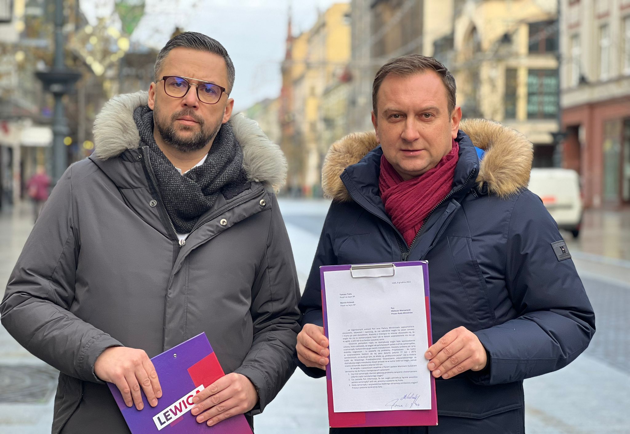 Dwóch mężczyzn w zimowych kurtkach z futrzanymi kapturami stoi na ulicy, trzymając foldery z dokumentami, z widocznym napisem Lewica.