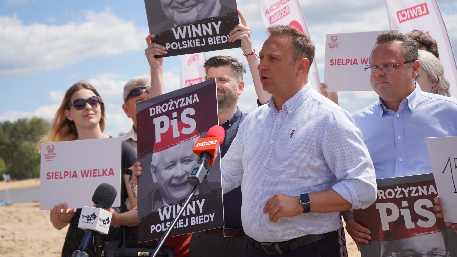 Na zdjęciu widać grupę osób trzymających transparenty i plakaty podczas protestu, z mężczyzną przemawiającym do mikrofonu.