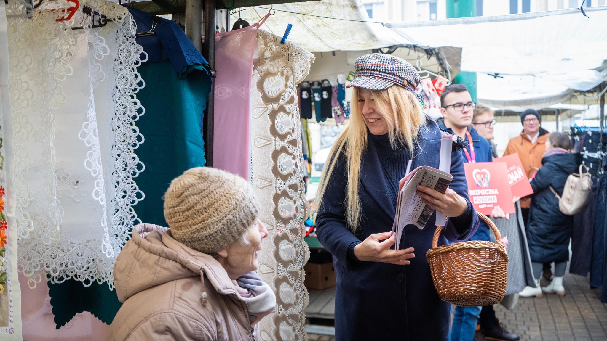 Uśmiechnięta kobieta z koszykiem w dłoni rozmawia z seniorką w beżowej czapce przy stoisku z koronkami i tkaninami na targu.