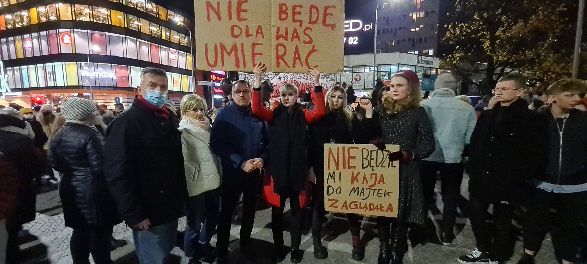 Grupa protestujących na wieczornym placu miasta trzyma kartonowe plakaty z hasłami antyprzemocowymi, a jedna osoba unosi duży plakat nad głową, w tle widać nowoczesne budynki.