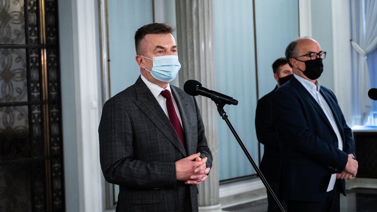 Dwóch mężczyzn w garniturach i maseczkach stoi przy mikrofonach podczas konferencji prasowej w eleganckim wnętrzu.