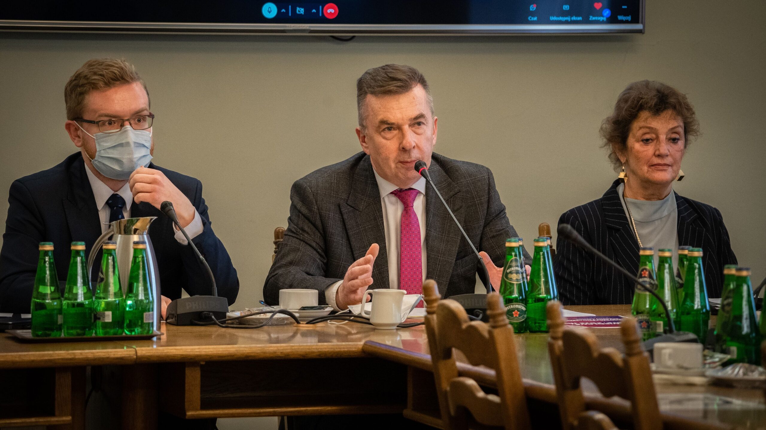 Na zdjęciu przedstawiono trzech uczestników spotkania przy stole, w tym mężczyznę przemawiającego z mikrofonem, obok którego siedzą dwie osoby, a na stole widoczne są zielone butelki wody.