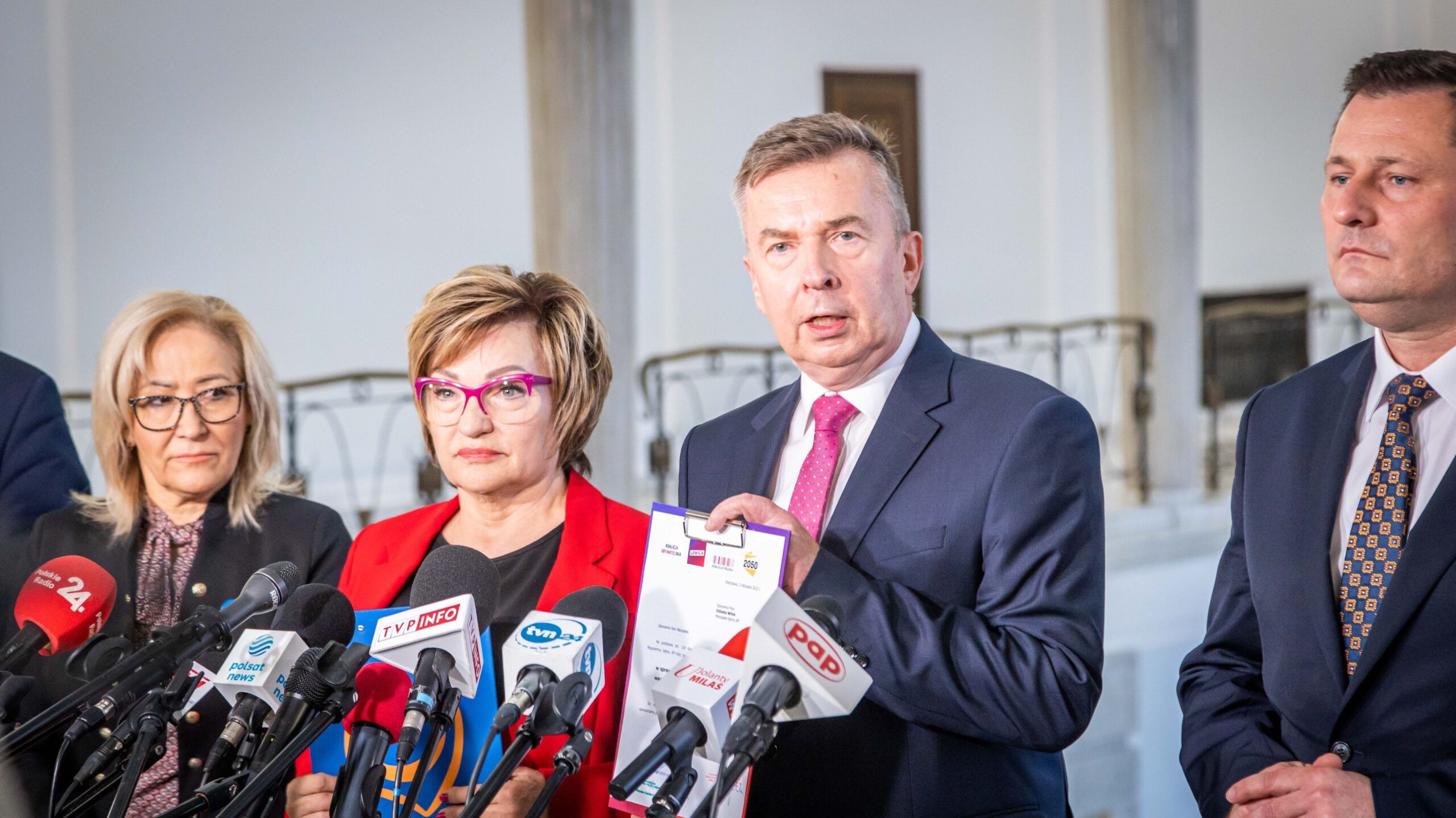 Na zdjęciu grupa polityków podczas konferencji prasowej, trzymających dokumenty i otoczonych mikrofonami w eleganckim wnętrzu.