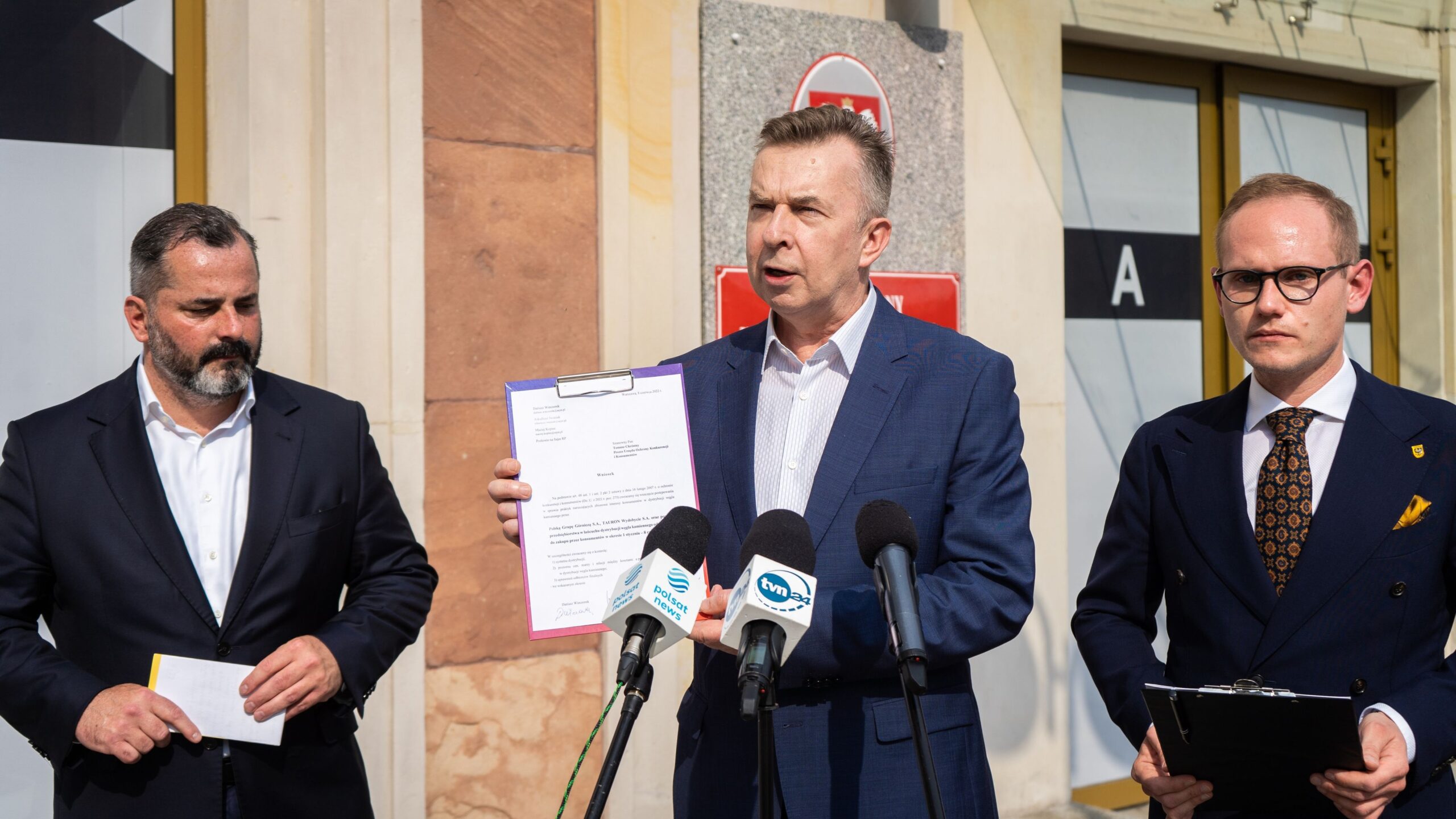Na zdjęciu widać trzech mężczyzn podczas konferencji prasowej, z jednym z nich trzymającym dokument, a w tle widoczne są drzwi budynku.