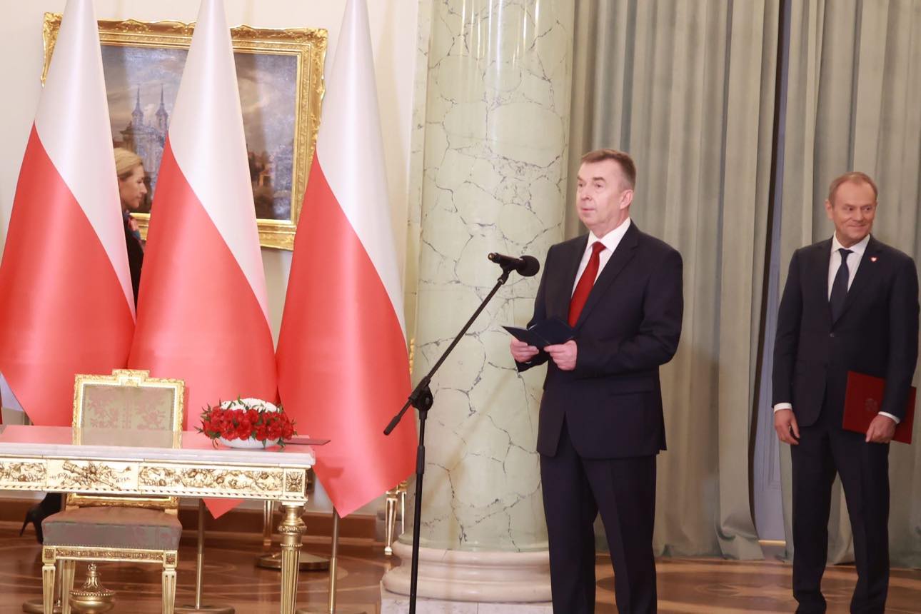 Mężczyzna w garniturze wygłasza przysięgę przy mikrofonie podczas zaprzysiężenia rządu, w tle stoją flagi Polski i ozdobna sala.