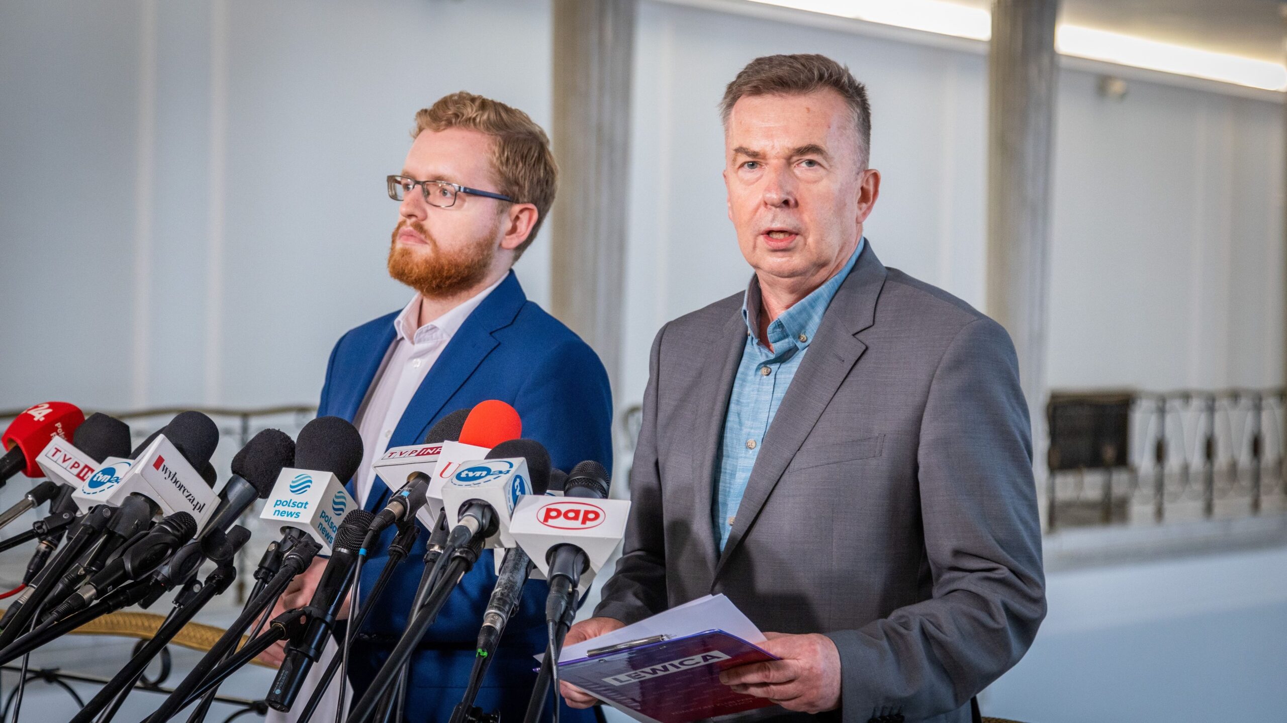 Dwóch polityków w garniturach stoi przy szeregu mikrofonów z logotypami mediów podczas konferencji prasowej dotyczącej rozbudowy elektrowni w Ostrołęce.