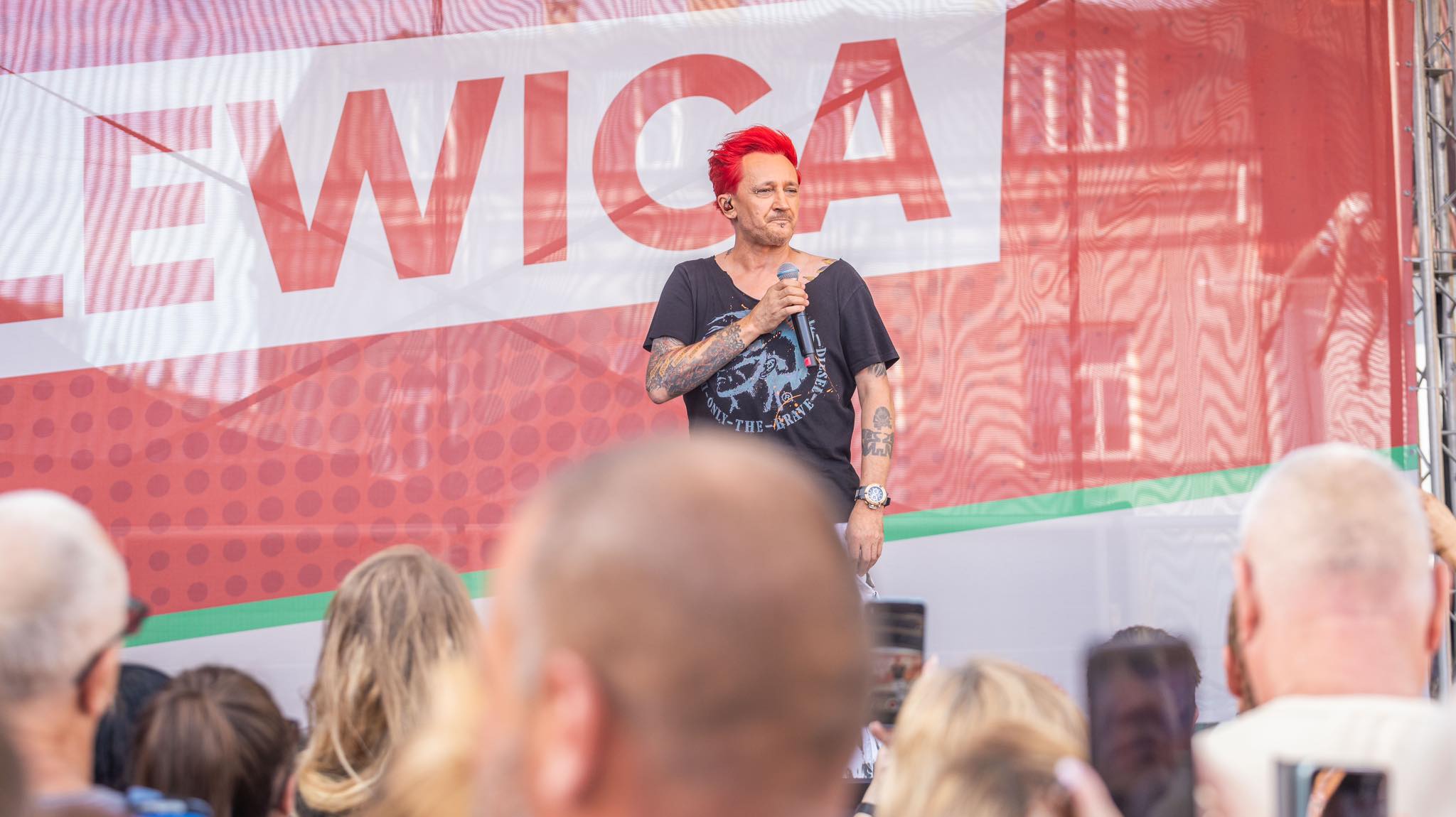 Osoba z czerwonymi włosami i tatuażami na ramionach stoi na scenie podczas wiecu w Częstochowie, trzyma mikrofon, a w tle widać duży baner z czerwonym napisem i zgromadzonych widzów.