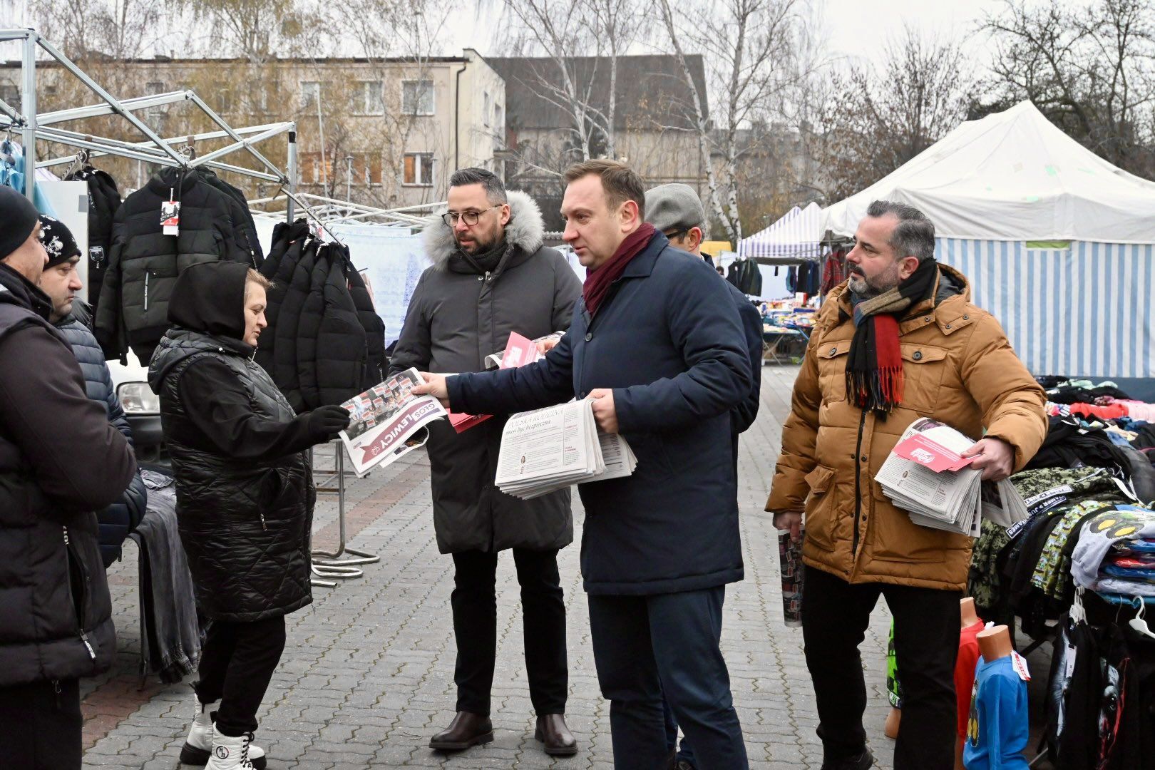Na zdjęciu grupa mężczyzn oraz jedna kobieta dyskutują na targu, gdzie sprzedawane są ubrania, a jeden z mężczyzn wręcza gazetę.