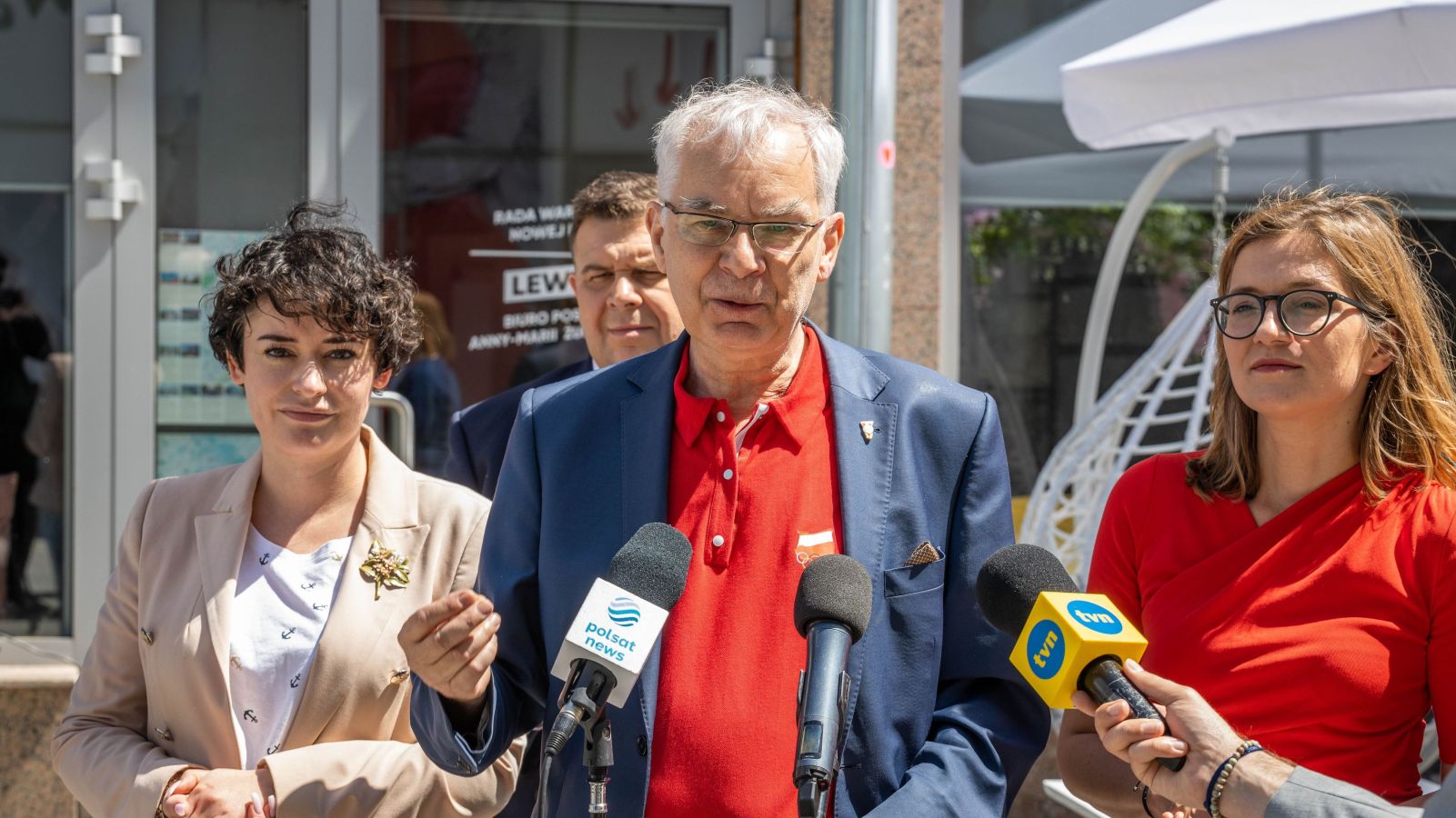 Grupa osób na zewnątrz podczas konferencji prasowej; mężczyzna w czerwonej koszuli i granatowej marynarce przemawia do mikrofonów, otoczony przez dwie kobiety, a na mikrofonach widać logotypy Polsat News i TVN.