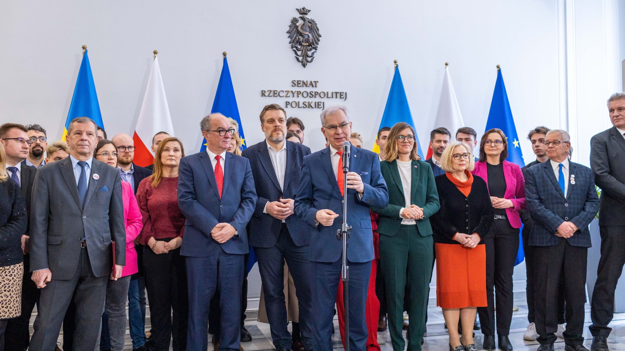 Grupa polityków Lewicy podczas konferencji prasowej, z centralnym mówcą przy mikrofonie, w tle flag Polski i Unii Europejskiej.