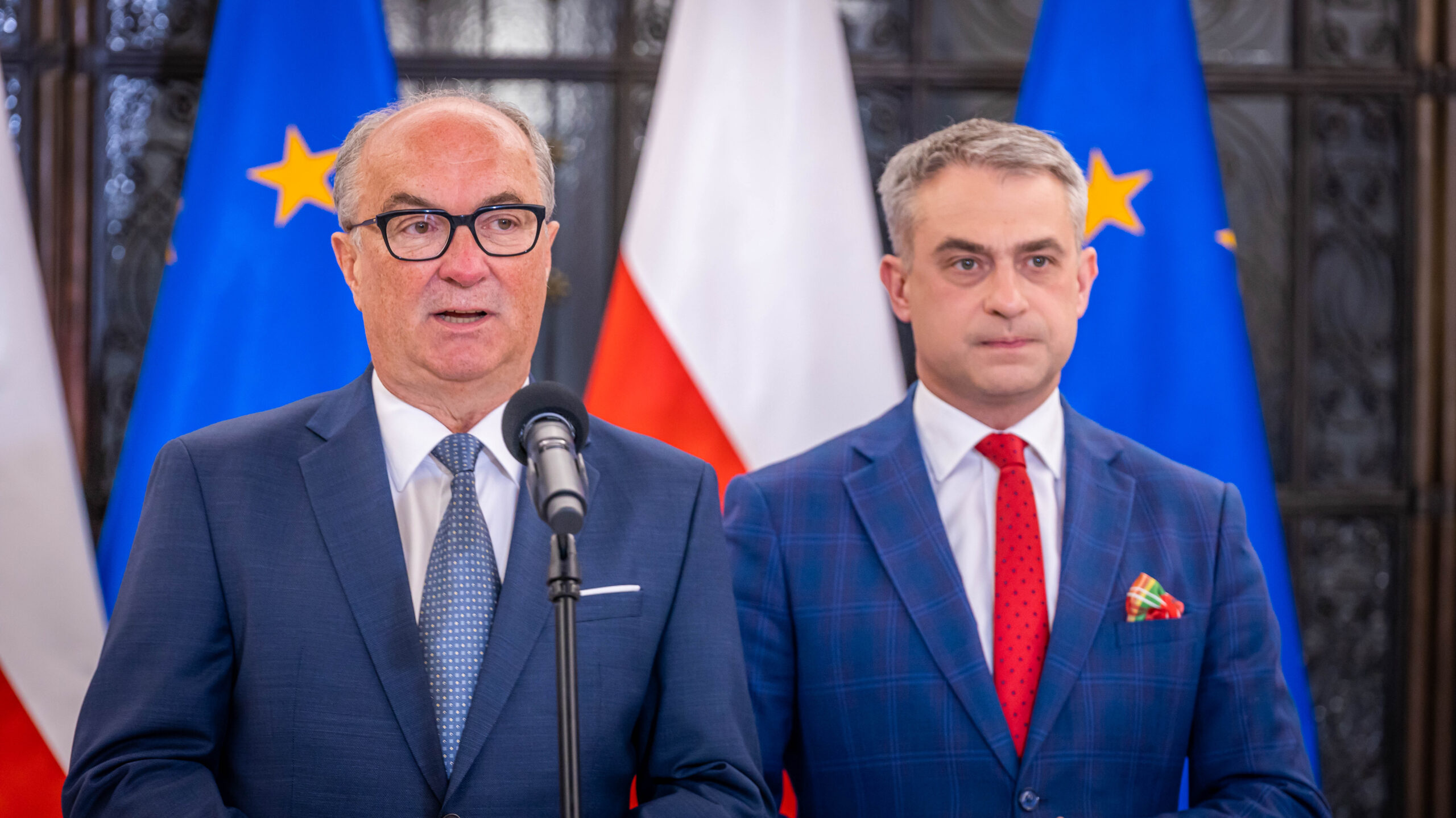 Dwóch mężczyzn w garniturach przemawia z mikrofonem podczas konferencji, na tle flag UE i Polski, symbolizując podpisanie Paktu Sejmowego i chęć współpracy oraz wysłuchania głosów społeczeństwa.