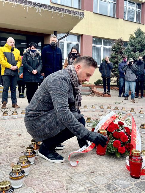 Na zdjęciu mężczyzna w szaliku składa kwiaty i zapala znicze w obecności innych osób podczas uroczystości pamięci.