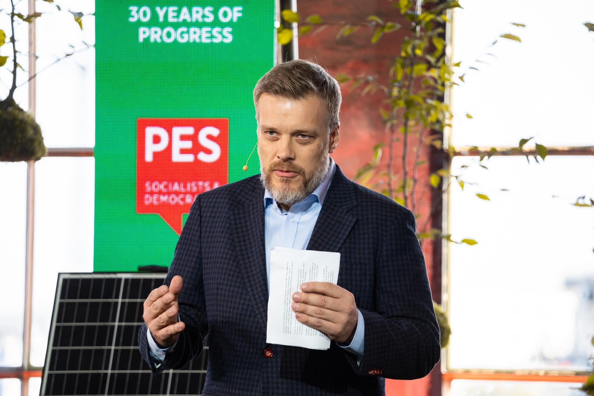 Mężczyzna w eleganckim ubraniu przemawia na tle kolorowego tła z logo PES, trzymając kartkę z notatkami.