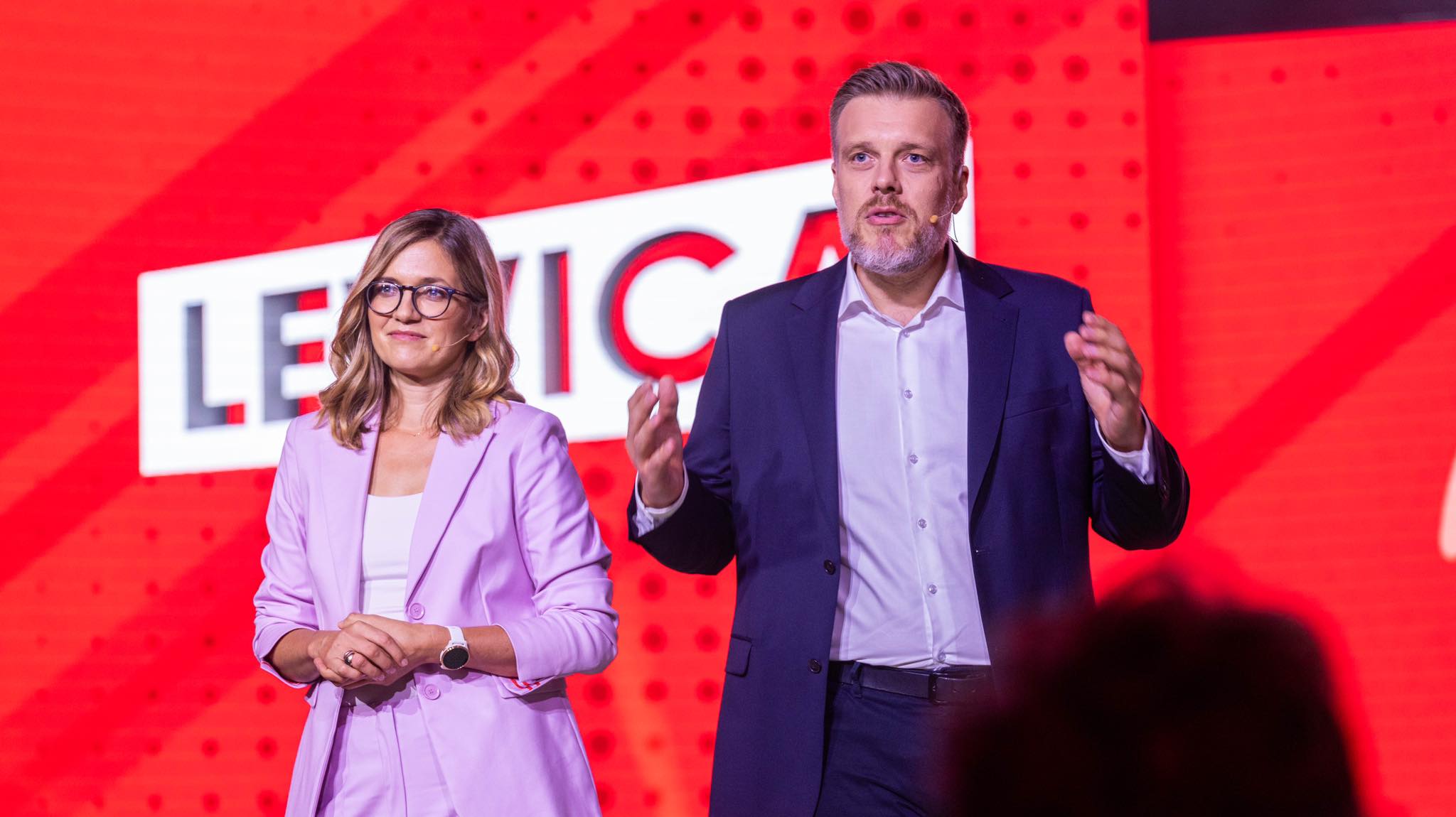Dwoje mówców na scenie podczas wydarzenia Lewicy: kobieta w jasnofioletowym garniturze i okularach po lewej, mężczyzna w granatowym garniturze po prawej, na czerwonym tle z napisem Lewica.