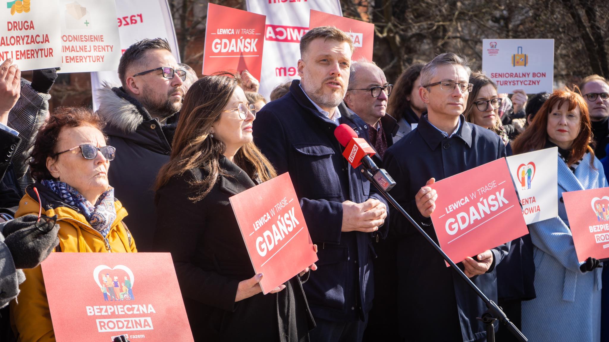 Grupa osób na wiecu politycznym trzyma czerwone plakaty z hasłem „Gdańsk” i innymi hasłami, podczas gdy mężczyzna przemawia do mikrofonu.