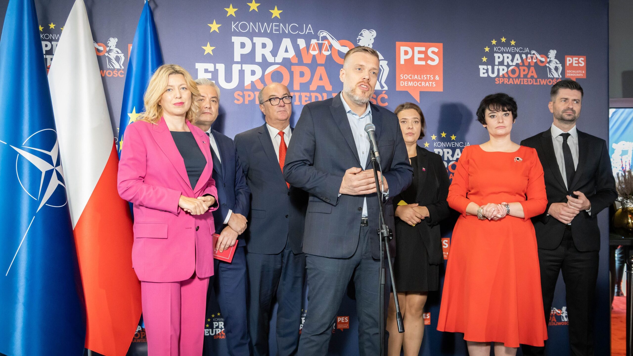 Na zdjęciu widać grupę osób stojących na scenie podczas konwencji, przedstawiających różne flagi i loga partii politycznych.