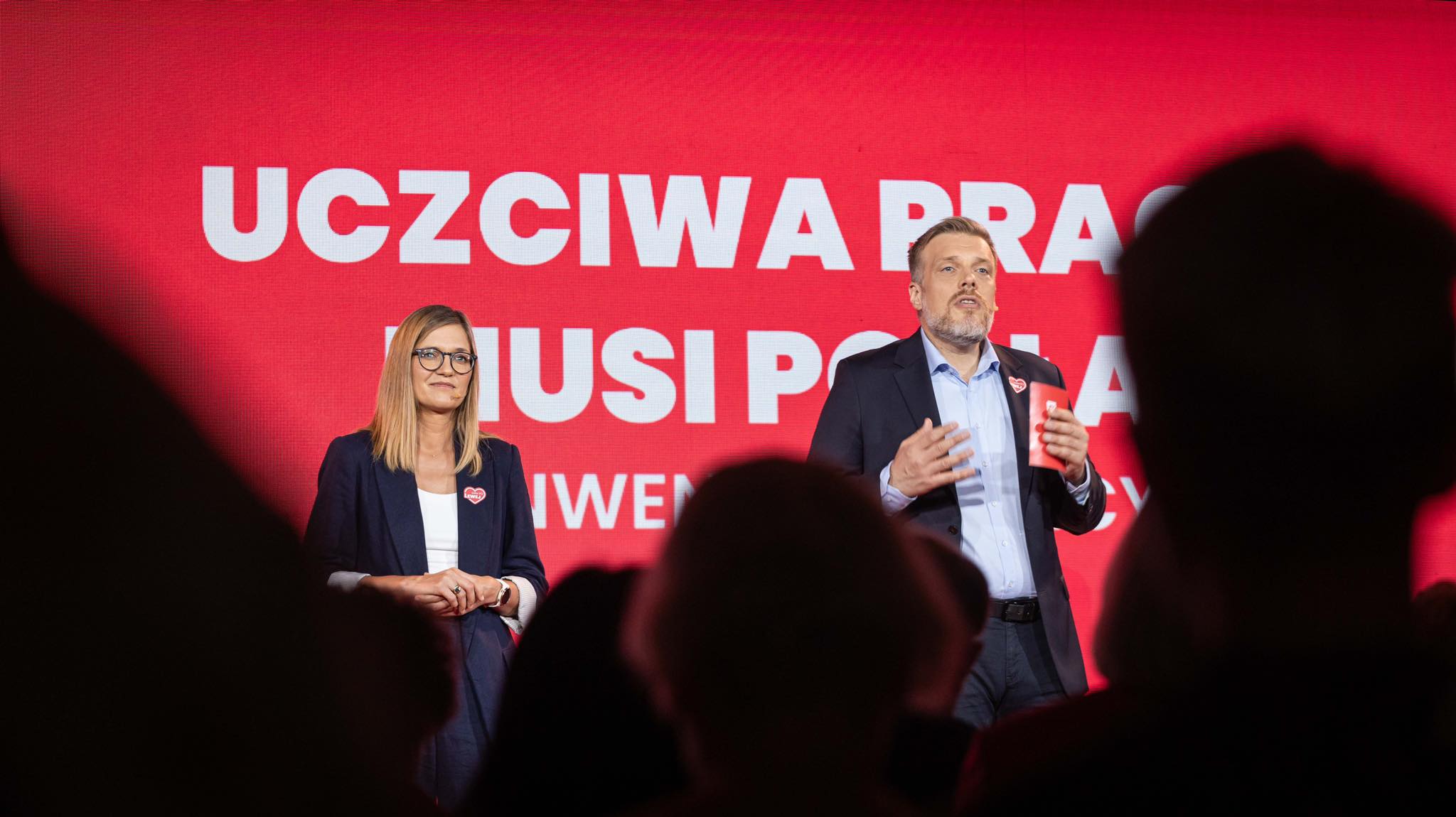 Dwoje mówców stoi na scenie na tle czerwonego baneru z dużym białym napisem „UCZCIWA PRACA MUSI PŁACAĆ”.