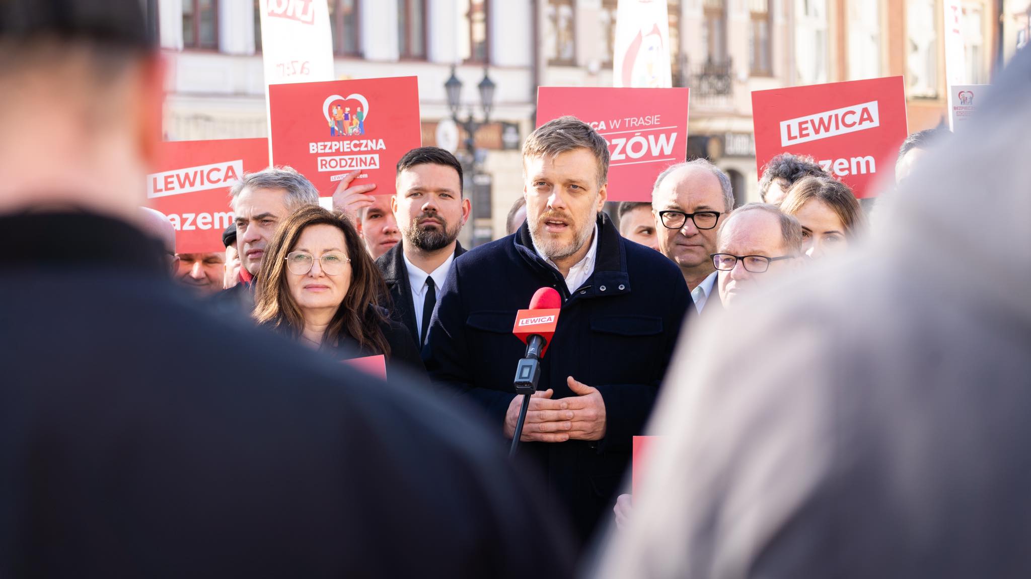 Na zdjęciu widoczna jest grupa osób stojących za mówcą podczas wydarzenia, trzymających czerwone plakaty z hasłami partii Lewica.