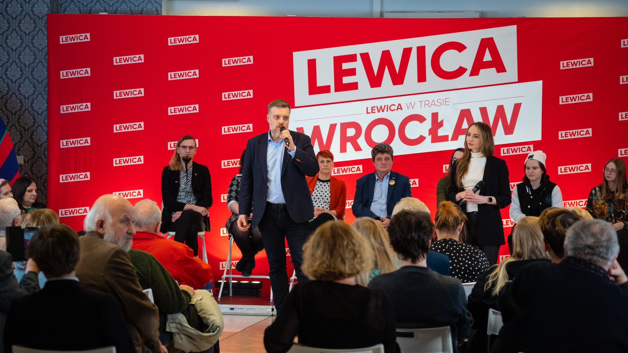 Na zdjęciu przedstawiona jest spotkanie partii Lewica we Wrocławiu, z mówcą na pierwszym planie i grupą uczestników siedzących w tle.