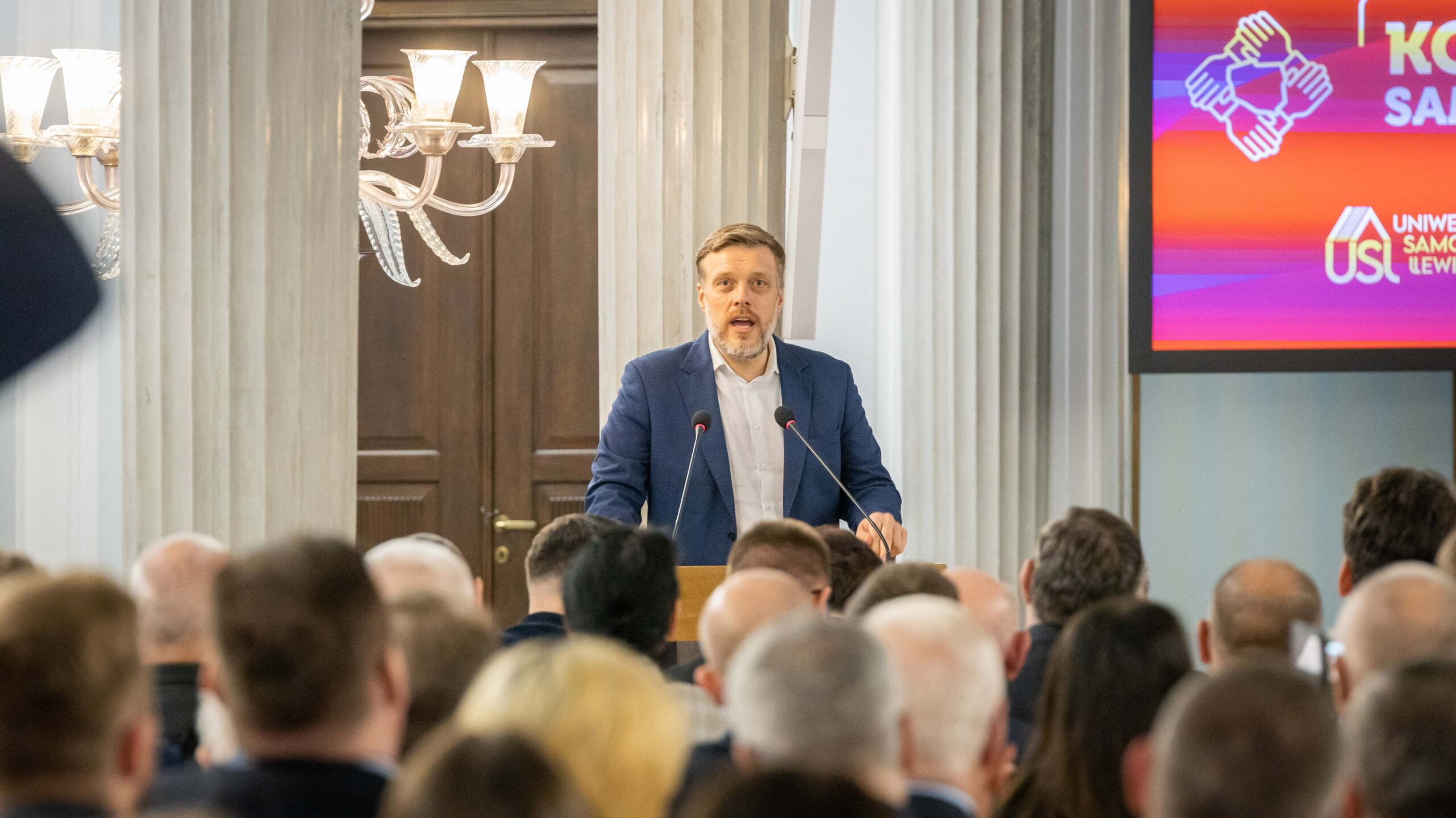 Na zdjęciu widoczny jest mężczyzna w niebieskim garniturze, przemawiający z mównicy podczas wystąpienia przed zgromadzonym tłumem w eleganckim wnętrzu.