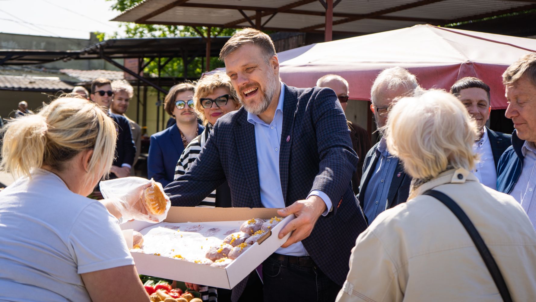 Uśmiechnięty mężczyzna w granatowej marynarce rozdaje wypieki z pudełka kobiecie w tle tłumu podczas wydarzenia Bezpieczna Rodzina w Kujawsko-Pomorskim.