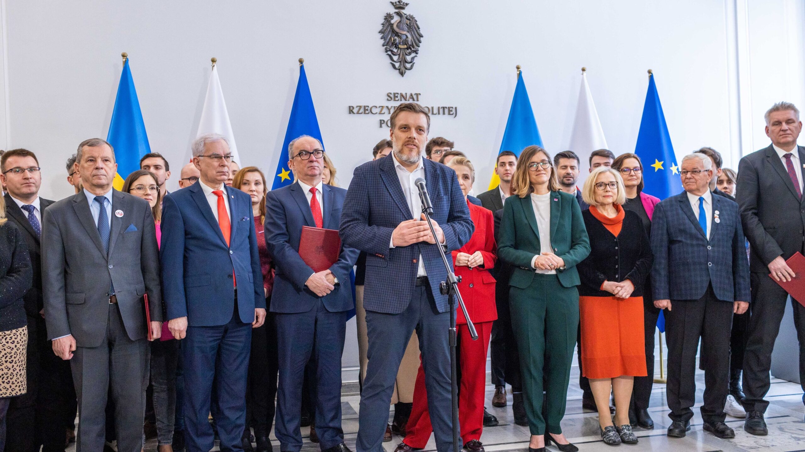 Grupa polityków w garniturach podczas konferencji prasowej w budynku Senatu RP, z centralnie stojącym mówcą przy mikrofonze i flagami w tle.