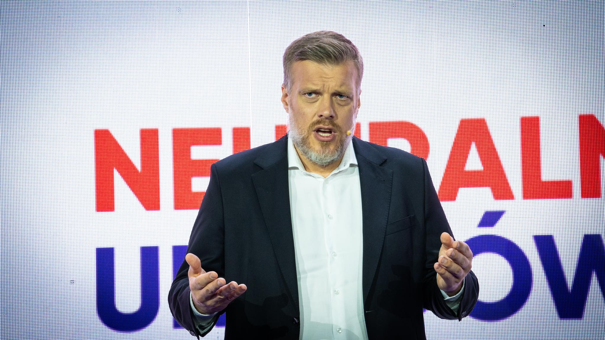 Mężczyzna w garniturze z mikrofonem na twarzy wygłasza przemówienie na scenie, na tle dużego ekranu z czerwonymi literami.