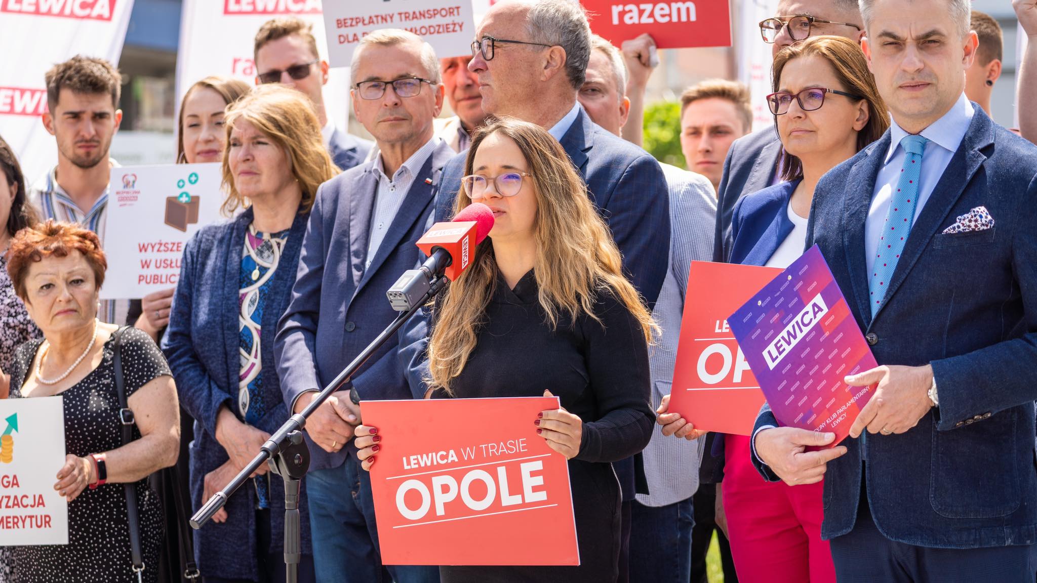 Kobieta z okularami przemawia do mikrofonu podczas wiecu Lewicy w Opolu, trzymając czerwony plakat „LEWICA W TRASIE OPOLE” wśród zgromadzonych i plakatów w tle.