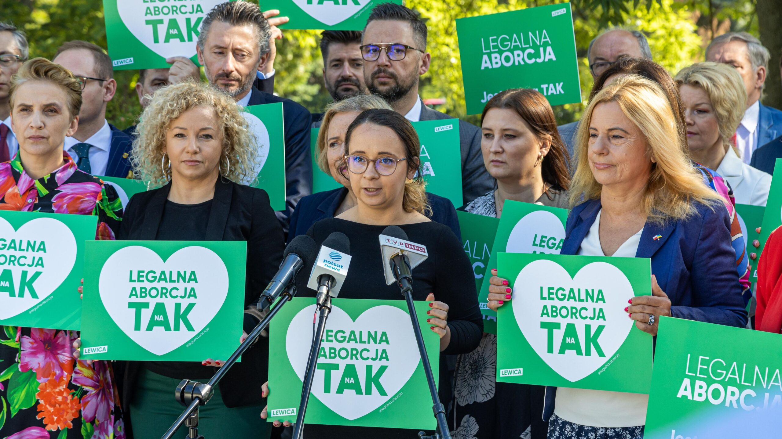 Na zdjęciu widoczna jest grupa osób trzymających zielone plakaty z napisem „LEGALNA ABORCJA TAK”, a jedna z kobiet przemawia do mikrofonu.