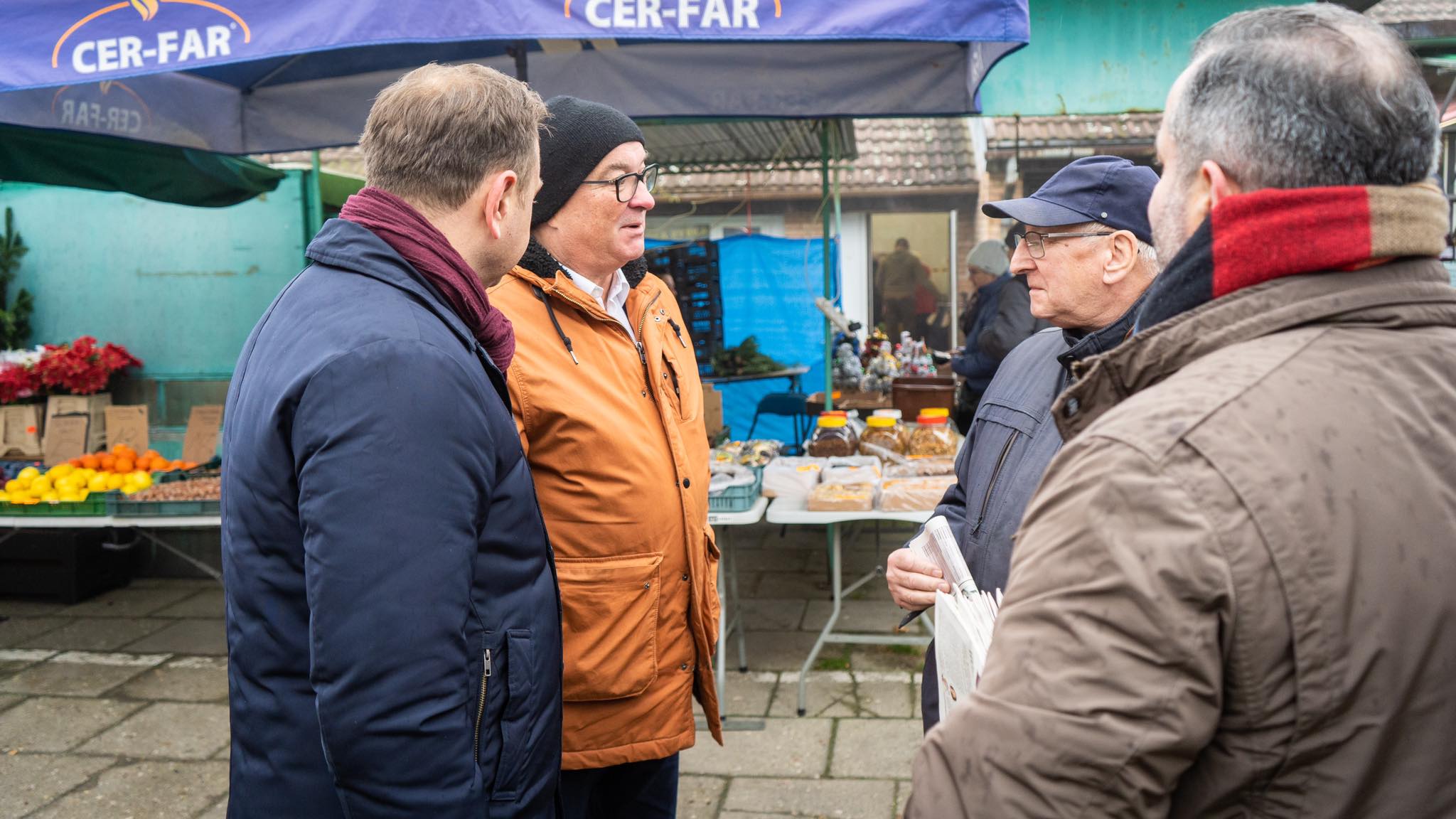 Grupa mężczyzn w zimowych kurtkach rozmawia na targowisku, stojąc przy stoisku z warzywami, słoikami i wypiekami pod niebieskim daszkiem CER-FAR.