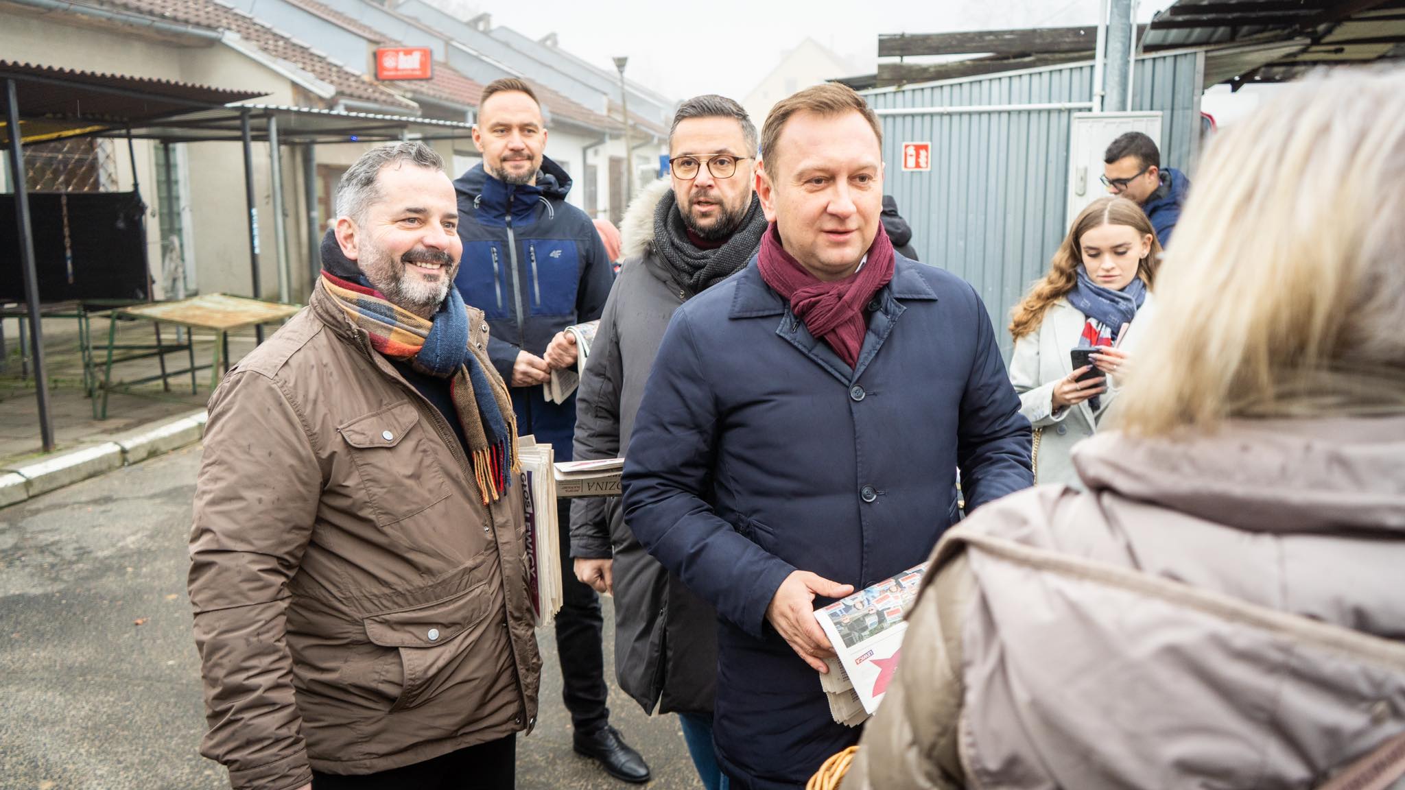 Grupa ludzi w zimowych kurtkach i szalikach stoi na ulicy podczas akcji Lewica w trasie, rozdaje gazety i rozmawia z przechodniami.