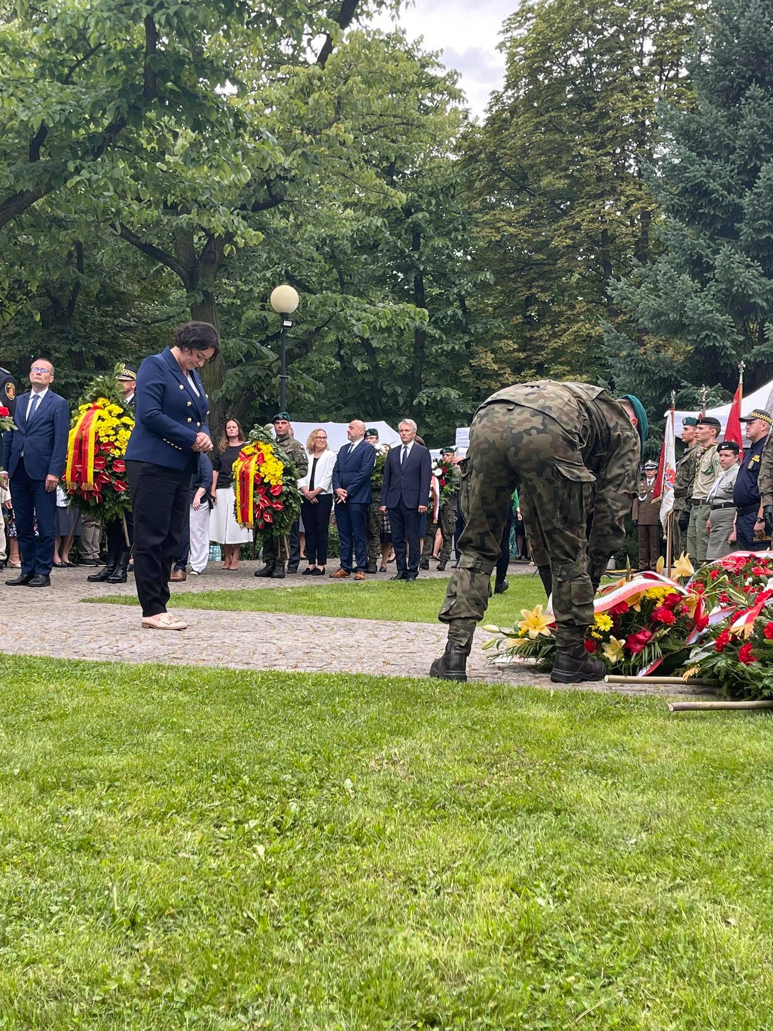 Grupa osób w garniturach i mundurach składa wieńce podczas ceremonii upamiętniającej Powstanie Warszawskie, a żołnierz w kamuflażu pochyla się nad kwiatami przy dekoracjach.