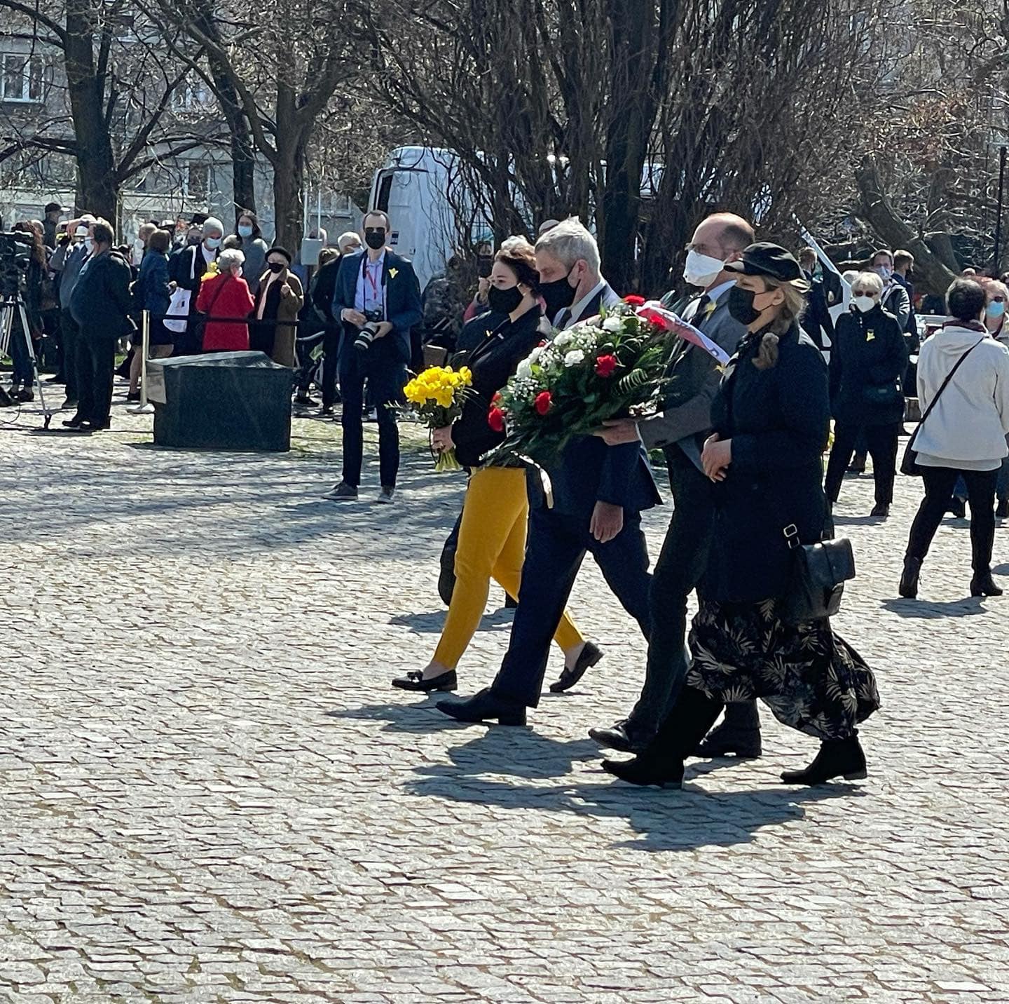 Grupa uczestników uroczystości z maseczkami idzie przez brukowany plac, niosąc kwiaty podczas obchodów 78. rocznicy Powstania w Getcie Warszawskim.