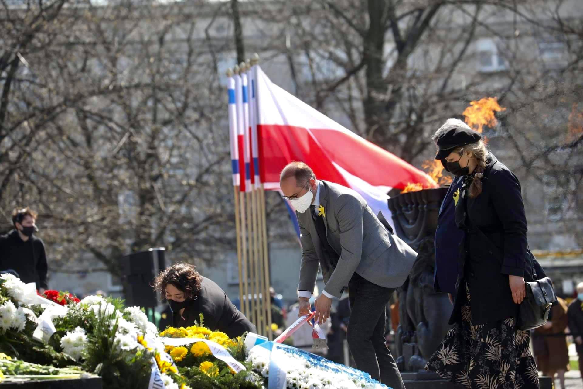 Uczestnicy obchodów 78. rocznicy Powstania w Getcie Warszawskim składają kwiaty przy płonącym ognisku, z polską flagą w tle.