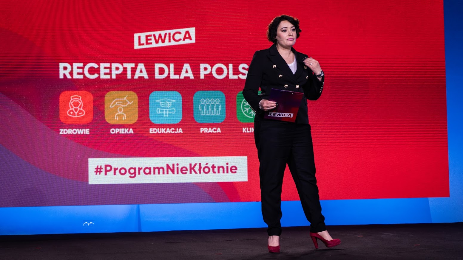 Kobieta w czarnym garniturze stoi na scenie przed czerwonym tłem z napisem Lewica i Recepta dla Polski, trzyma teczkę i ma czerwone szpilki.