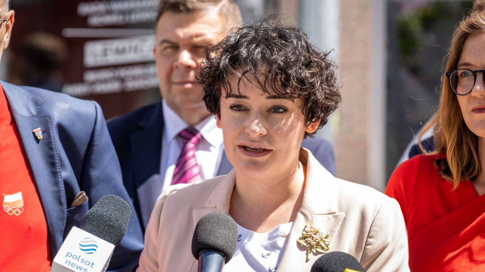 Kobieta z krótkimi czarnymi włosami w beżowym żakiecie z ozdobą na klapie przemawia do mikrofonów podczas konferencji prasowej na zewnątrz, w kontekście Dnia Dziecka.